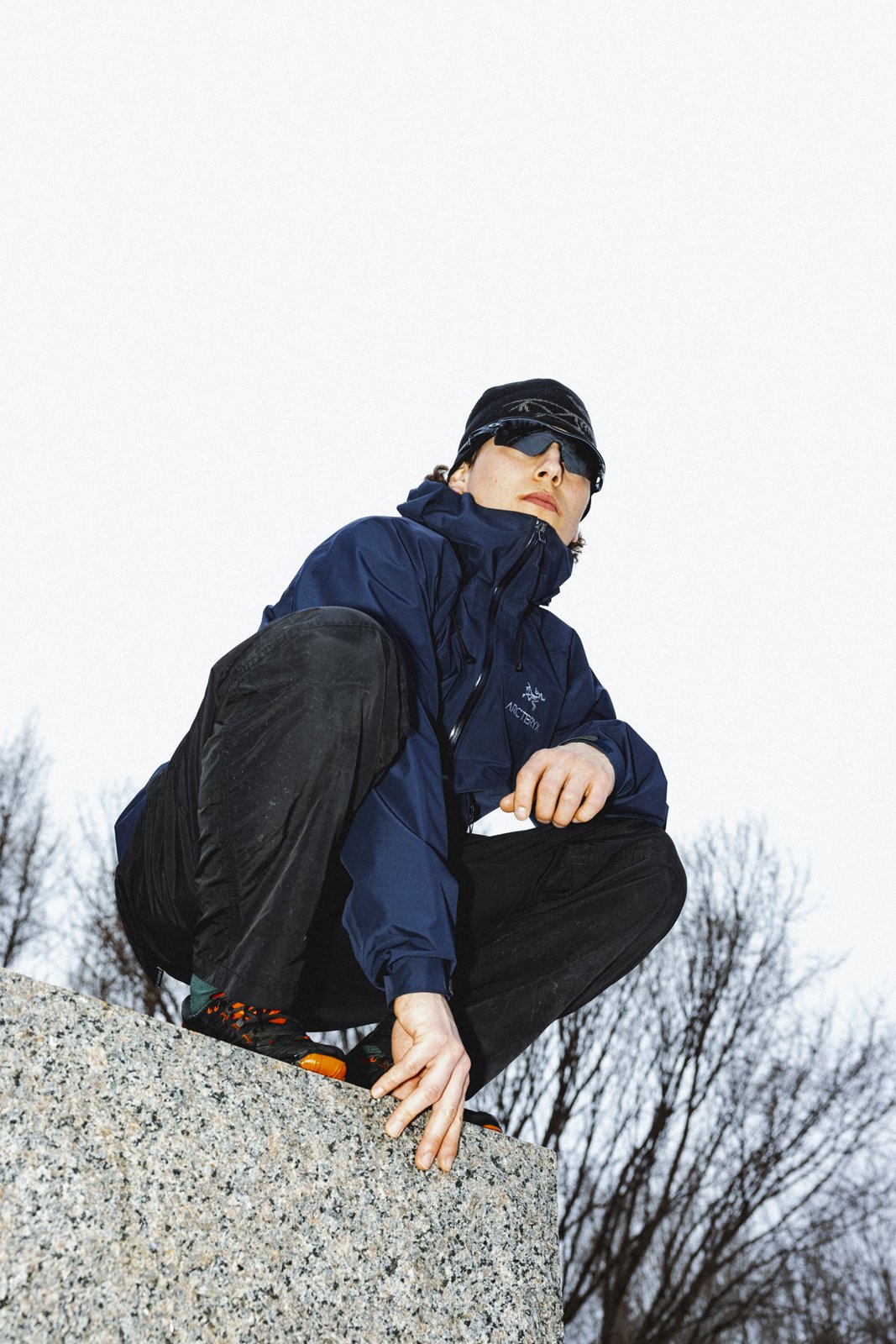 Homme en vêtements de randonnée, portant une veste bleue, un bonnet noir, des lunettes de soleil, pose sur une roche, arrière-plan de ciel gris et d'arbres dénudés.
