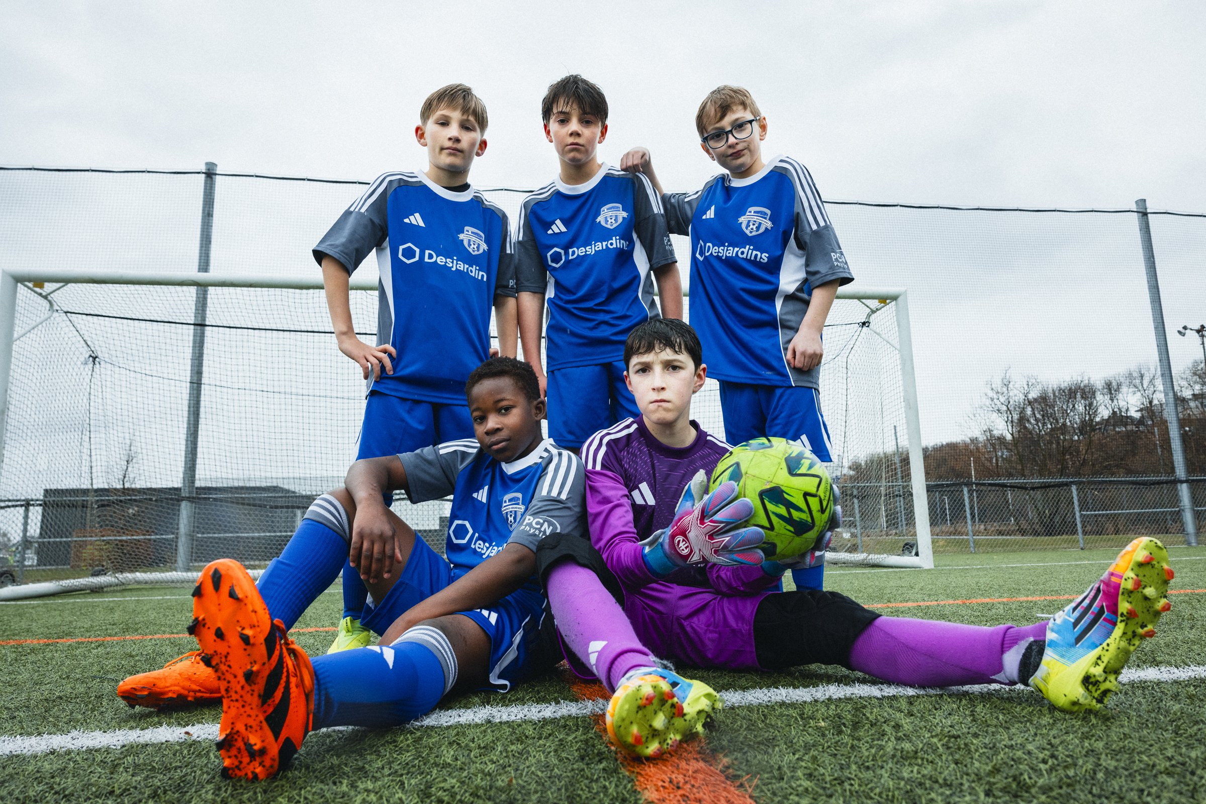 Groupe de cinq jeunes joueurs de football sur un terrain en synthétique, avec deux en position assise au premier plan et trois debout à l'arrière, portant des maillots bleus, une balise verte de football, et un ballon dans les mains d'un gardien en t