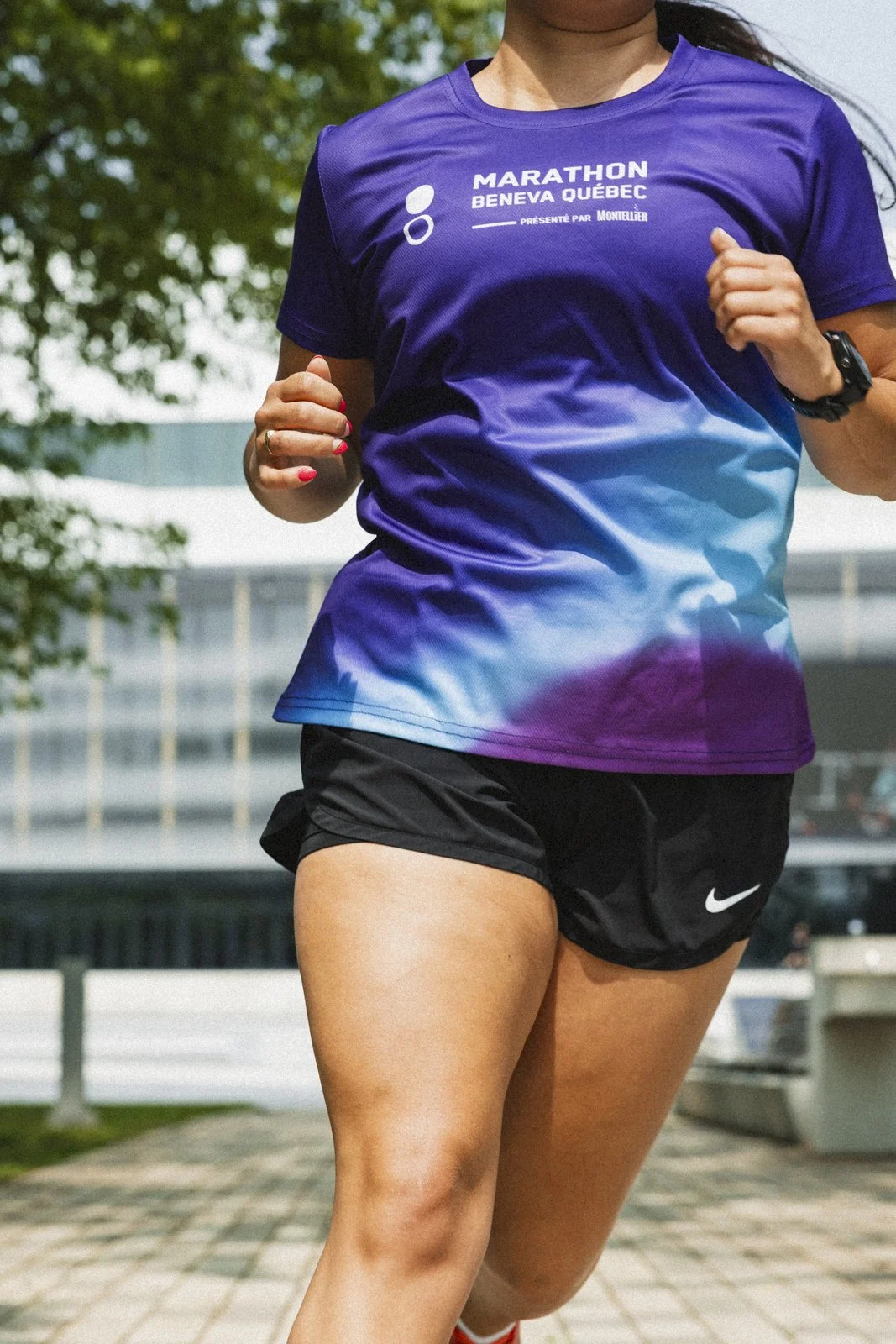 Profil d'une femme en tenue de course, portant un t-shirt violet avec l'inscription 'Marathon Beneva Québec' et un short noir Nike, courant en extérieur.