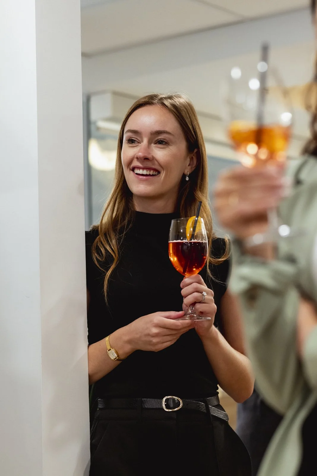Femme souriante tenant un verre de cocktail rouge avec une tranche d'orange, lors d'une fête ou un rassemblement social.