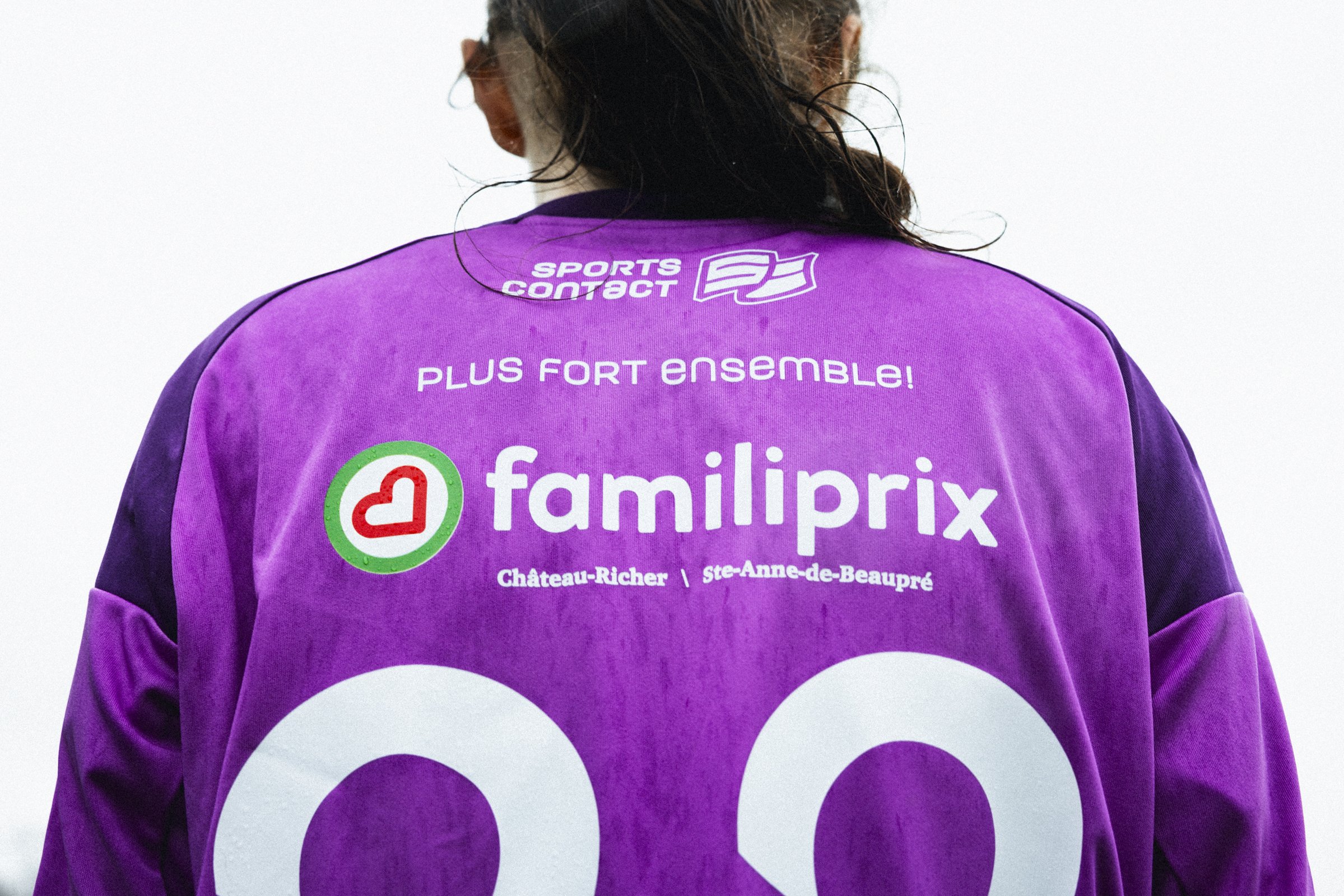 Vue de dos d'une personne portant un maillot de sport violet avec des logos, notamment 'fmpiliprix' et 'SPORTS CONTACT', sur une surface blanche.
