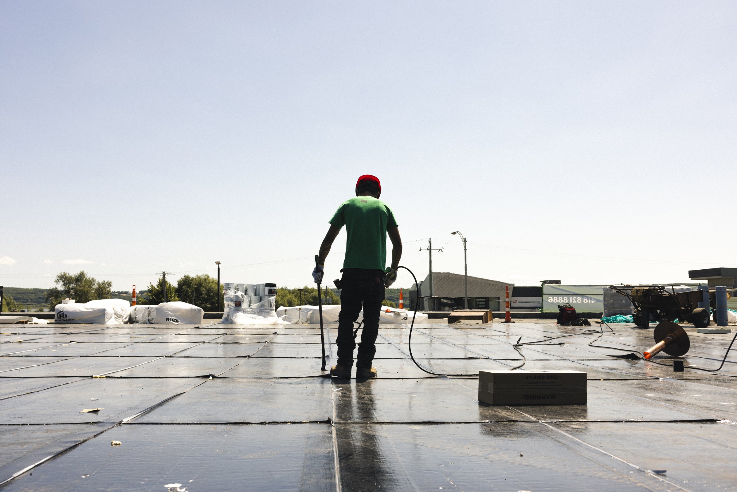 Travailleur sur un toit solaire, portant un casque rouge et des outils, sous un ciel clair.
