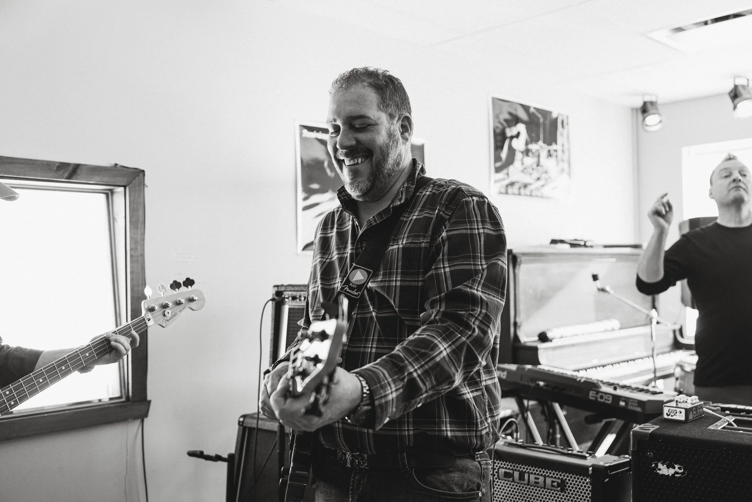Groupe de musiciens jouant dans une salle de répétition, un homme au centre joue de la guitare électrique, un autre homme à droite danse ou chante, tous deux sont impliqués dans une session musicale