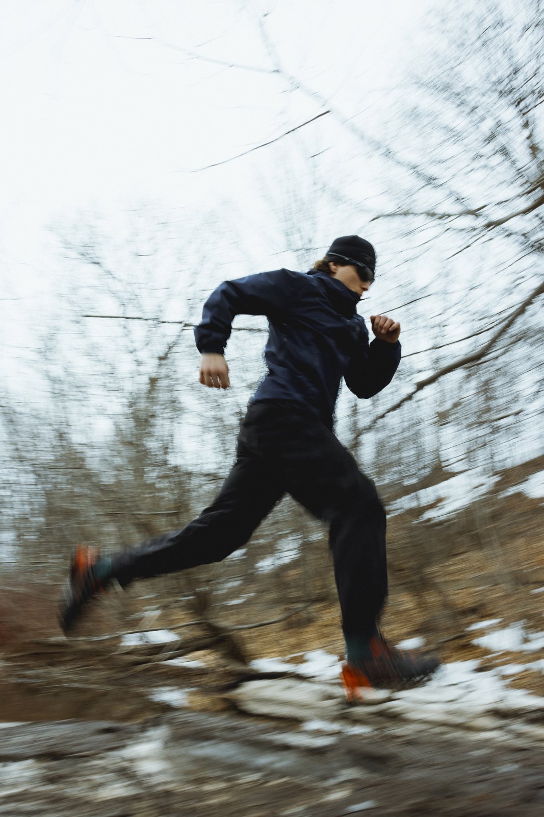 Un homme en vêtements de course, portant un bonnet et des lunettes de soleil, court rapidement à travers une forêt en hiver, avec des branches d'arbres défilant rapidement en arrière-plan.