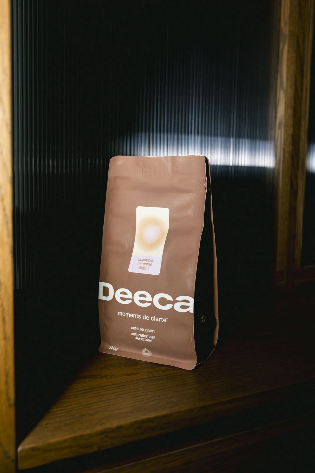 Un paquet de café en grains Deeca placé sur une étagère en bois, avec un fond sombre.