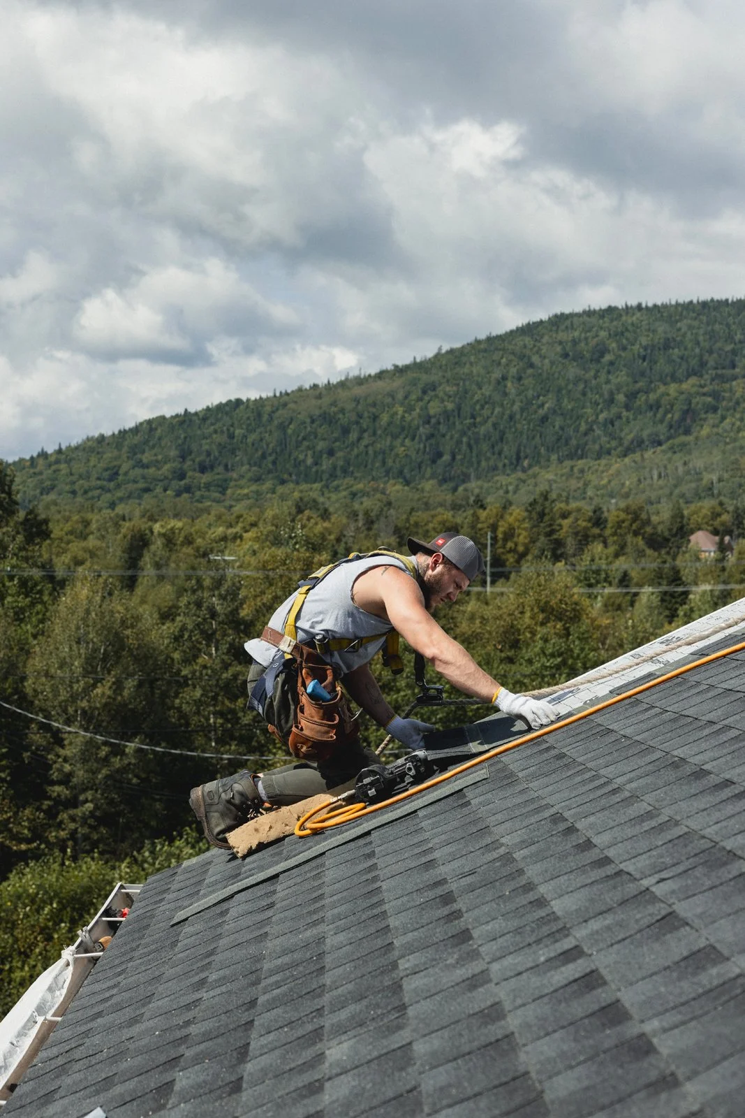 Ouvrier nettoyant ou réparant une toiture inclinée avec des outils et équipement de sécurité. En arrière-plan, une forêt et des collines sous un ciel nuageux.