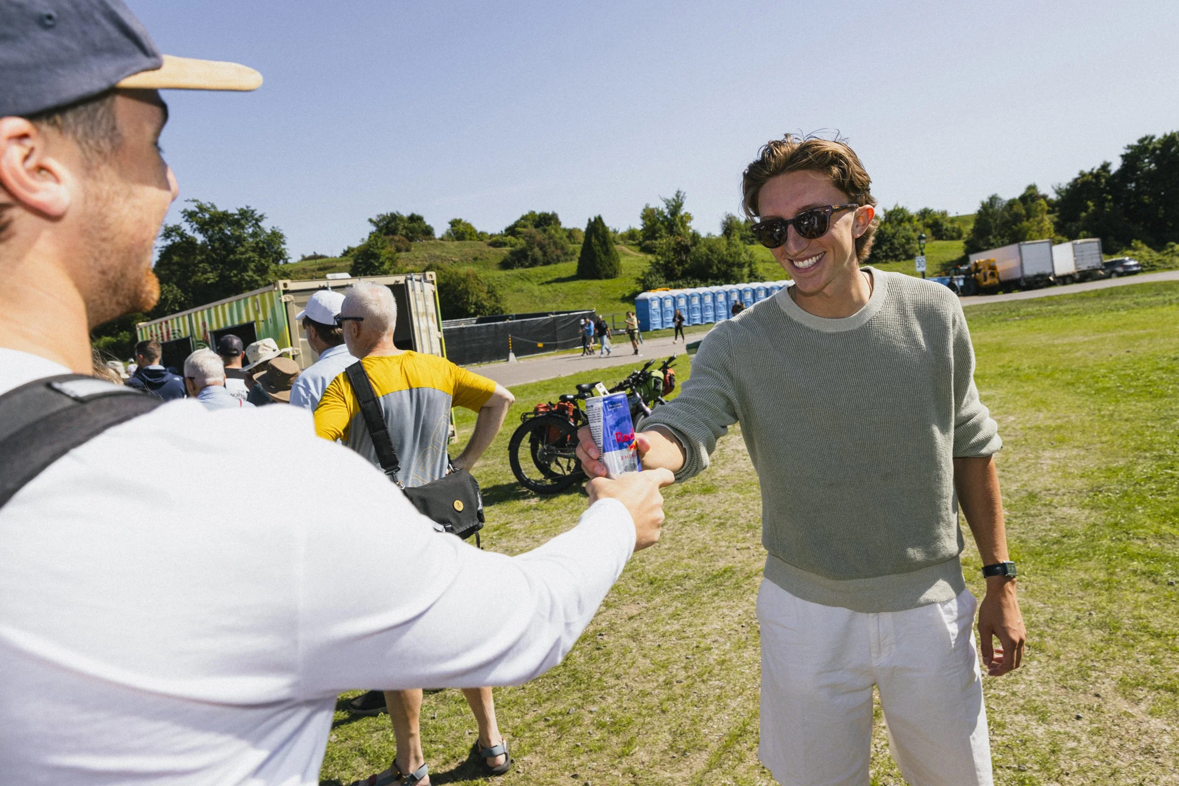 Un homme en chemise blanche donne une canette à un jeune homme en chemise gris clair, portant des lunettes de soleil, lors d'un événement en plein air sous un ciel ensoleillé.