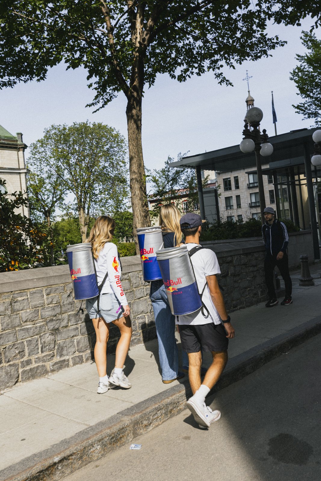 Trois personnes portant des sacs à dos Red Bull en forme de canette se promènent dans une rue urbaine, avec des bâtiments et des arbres en arrière-plan.