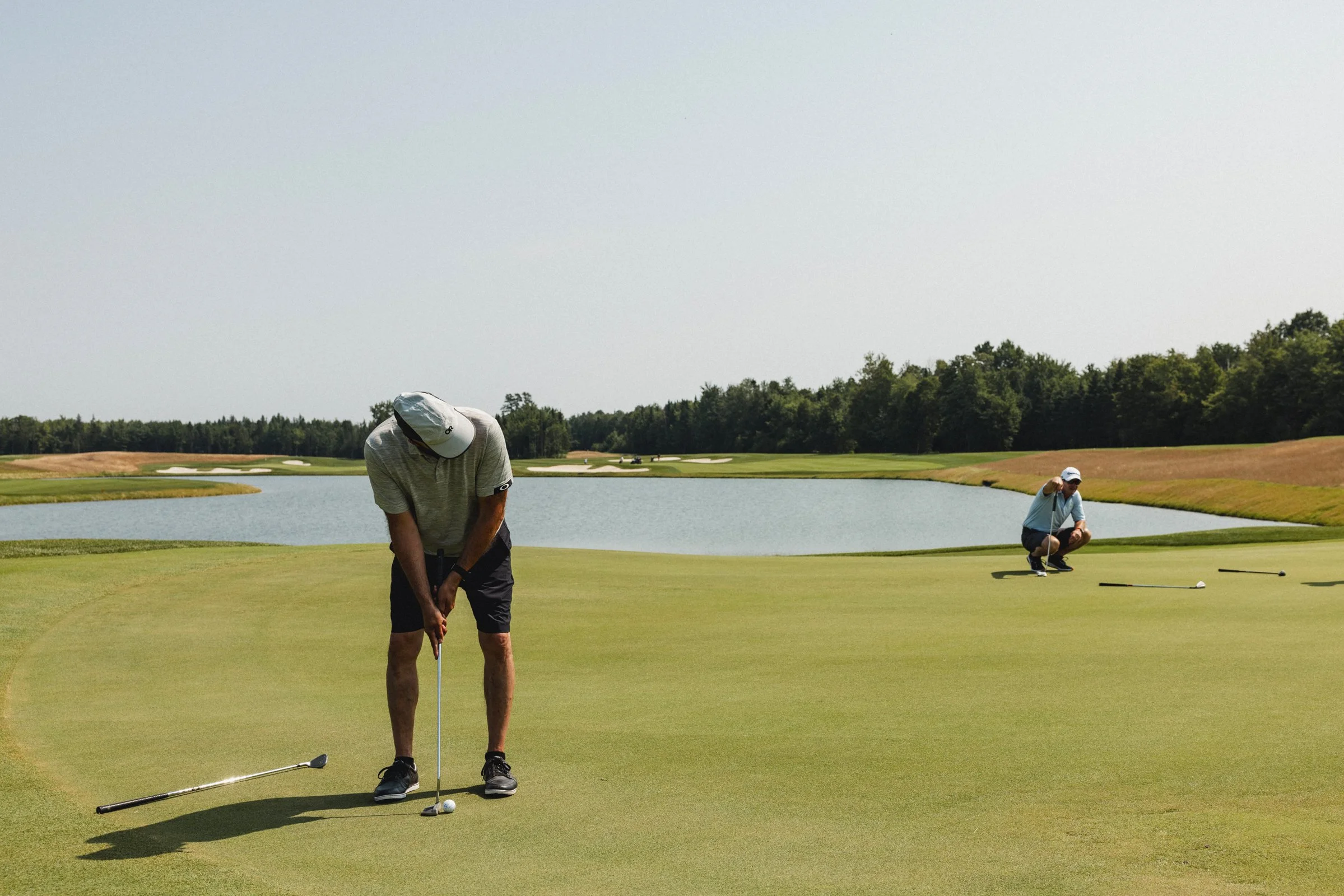 Deux hommes jouent au golf sur un terrain avec un étang en arrière-plan. L'un d'eux est en train de se préparer à frapper la balle, tandis que l'autre est accroupi, observant.