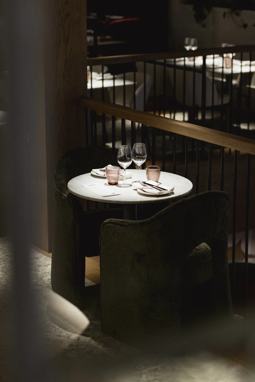 Une table élégante avec deux verres à vin, deux tasses en verre rose, des fourchettes, des couteaux, des assiettes et des serviettes placées dans un restaurant chic, avec des chaises vert foncé modernes et un décor intime.