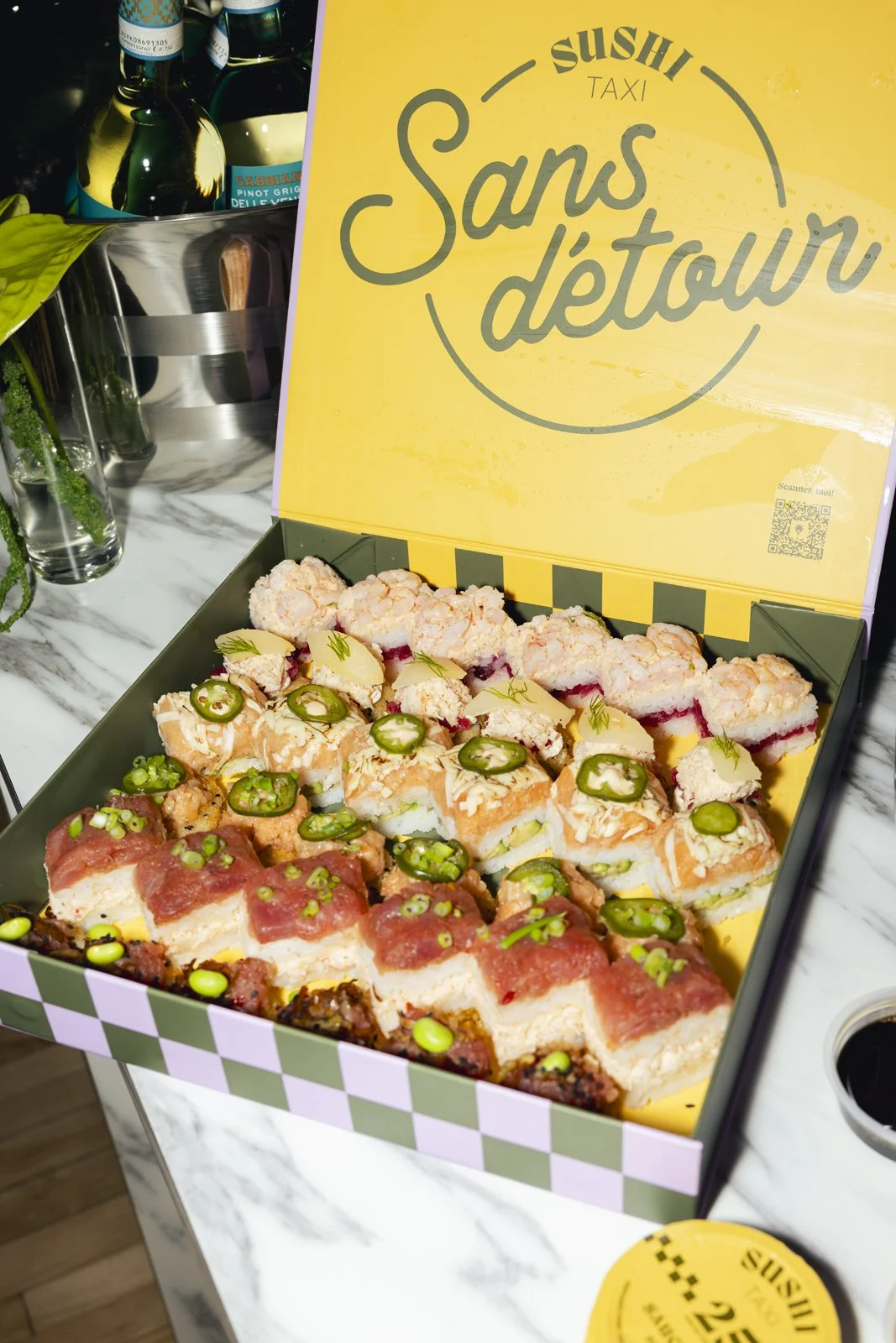 Une boîte contenant différents types de sushis décorés de jalapeños, de graines et d'autres garnitures, avec une couverture jaune portant l'inscription 'Sushi Taxi Sans d'town'.
