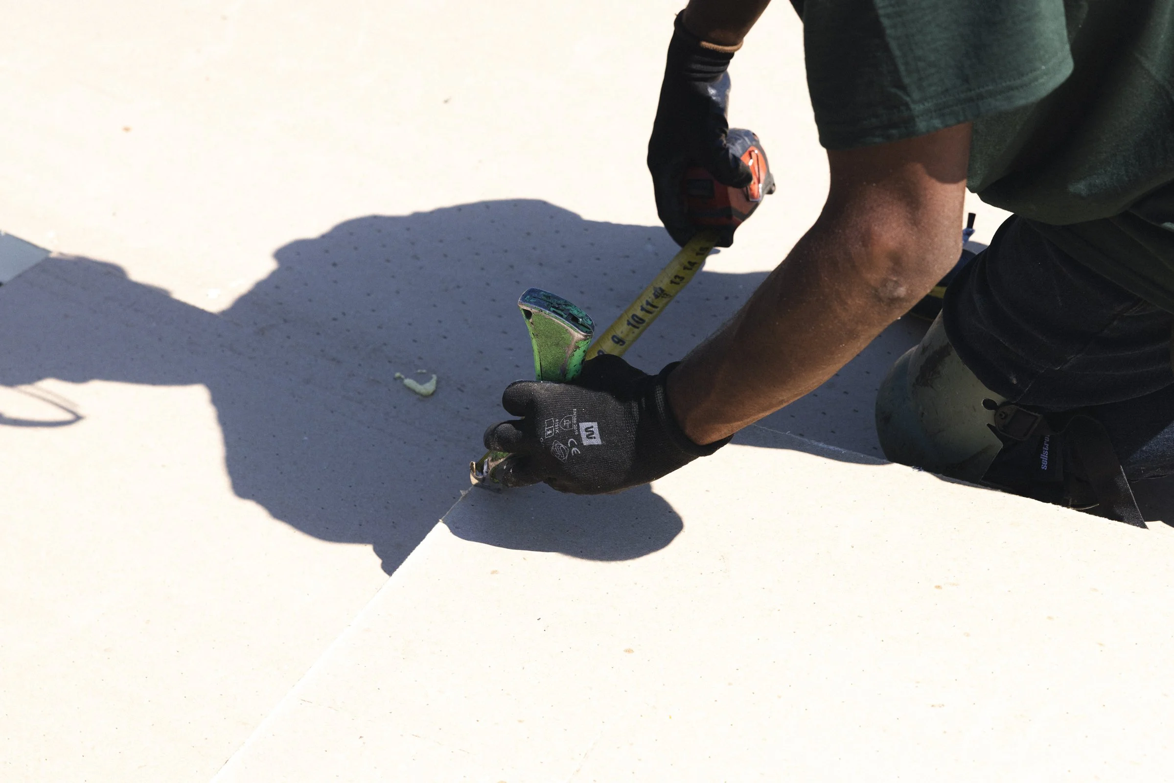 Une personne en train de faire des travaux, mesurant une distance à l'aide d'un ruban à mesurer et d'un marteau, portant des gants noirs et des vêtements de travail, posée sur une surface blanche ou claire.