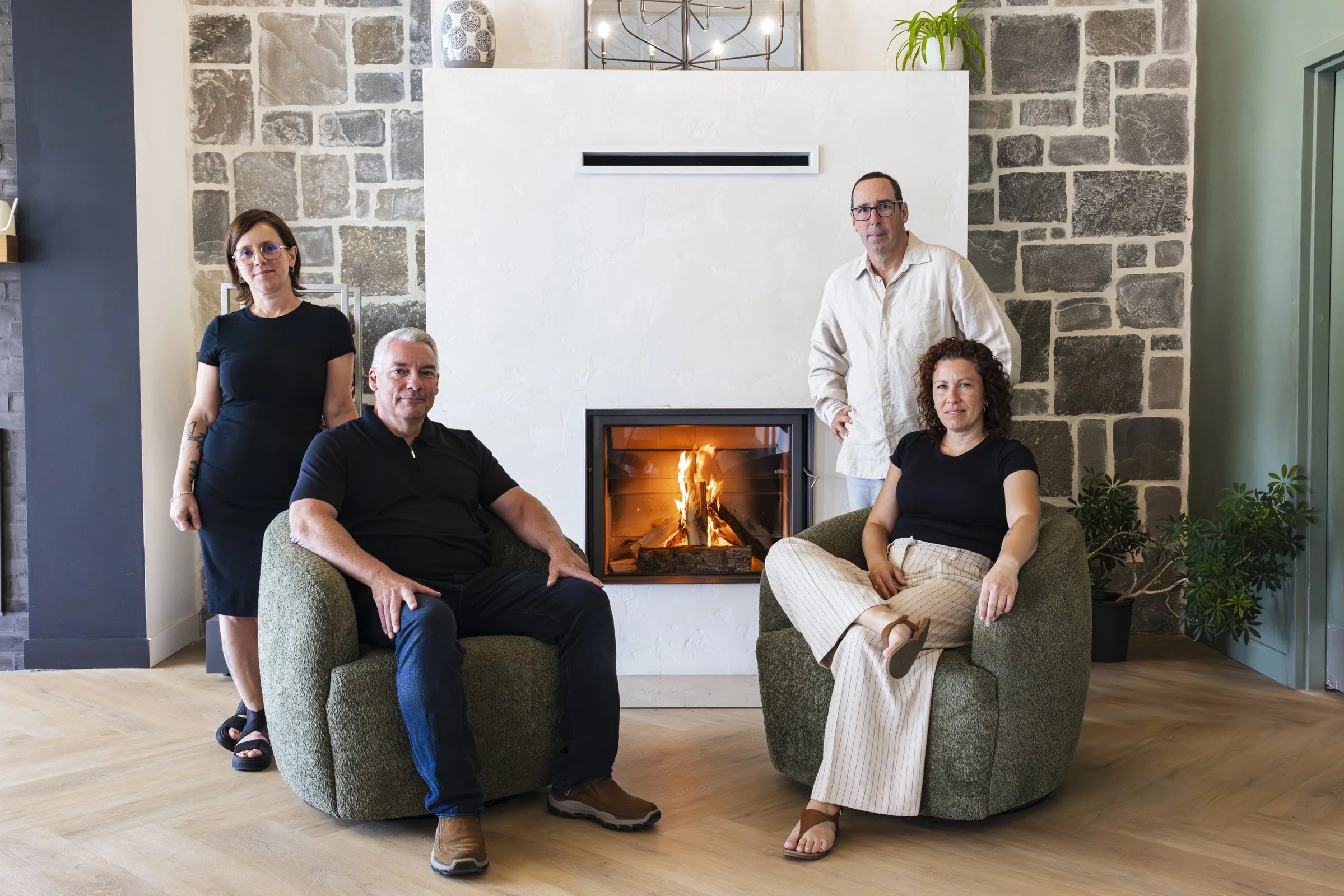 Cinq personnes posent dans un salon moderne avec cheminée allumée, deux assis dans des fauteuils verts et deux debout derrière, un homme et une femme. Le fond est constitué d'un mur en pierre grise et en blanc, avec des plantes décoratives.