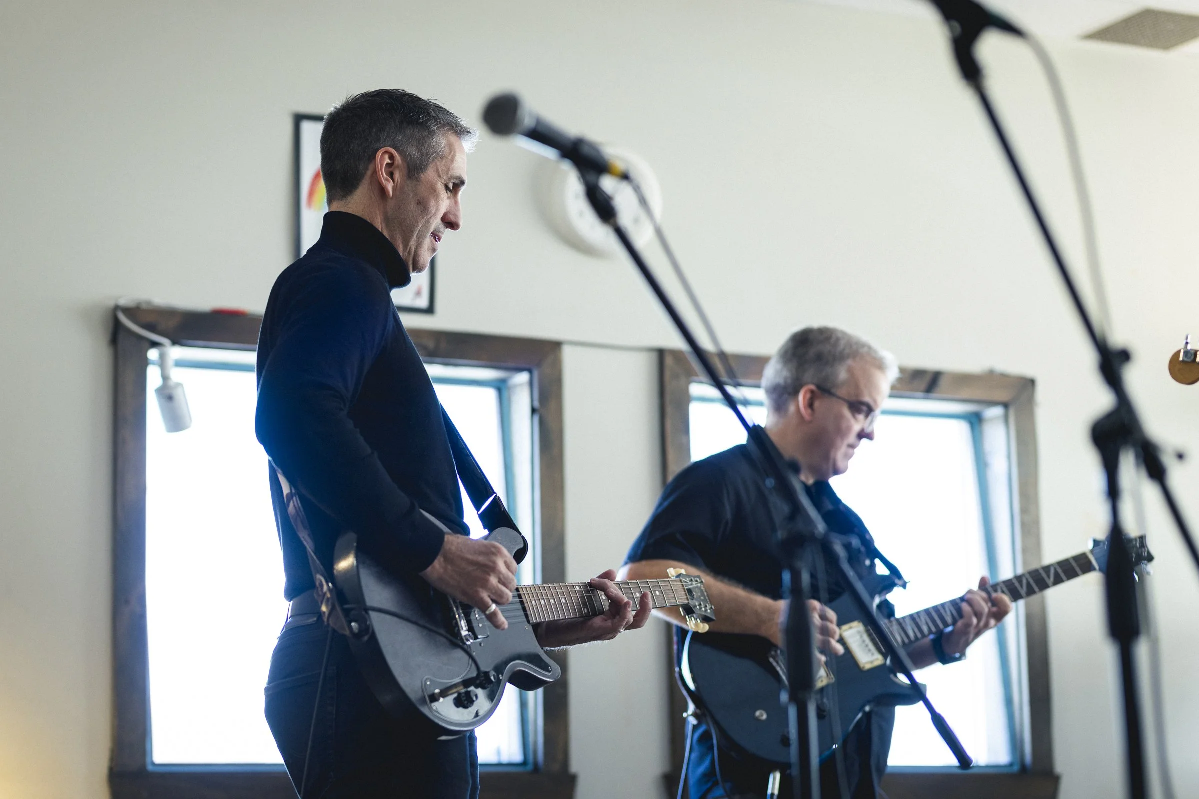 Deux musiciens jouent de la guitare lors d'une performance dans une pièce lumineuse avec deux fenêtres.}