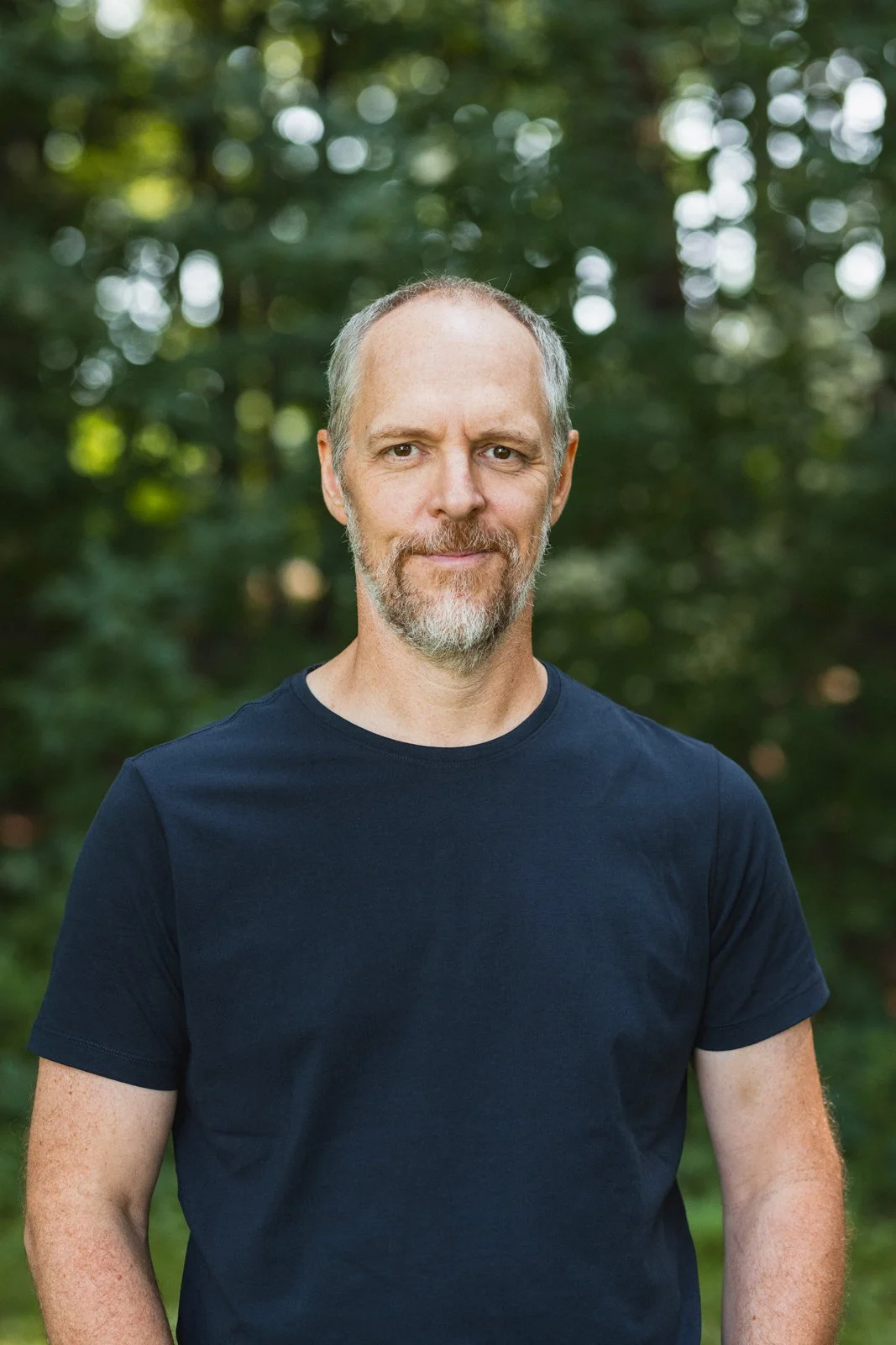 Un homme à la barbe et aux cheveux courts, portant un t-shirt noir, debout devant un fond de forêt verdoyante.