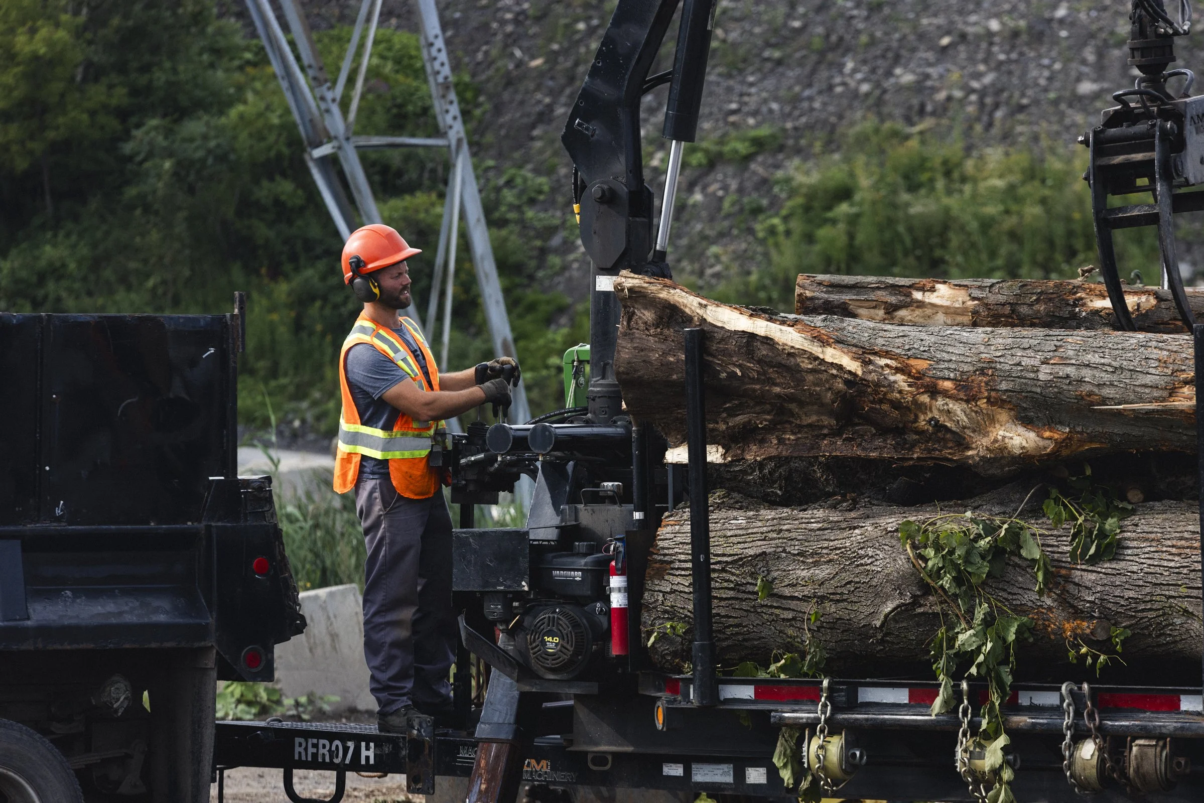 Un homme portant un casque orange, une veste de sécurité fluorescente et des écouteurs est à côté d'une grande table de coupe de bois. Il travaille avec une machine pour couper un gros troncs d'arbre sur une remorque attachée à un véhicule. Le fond m