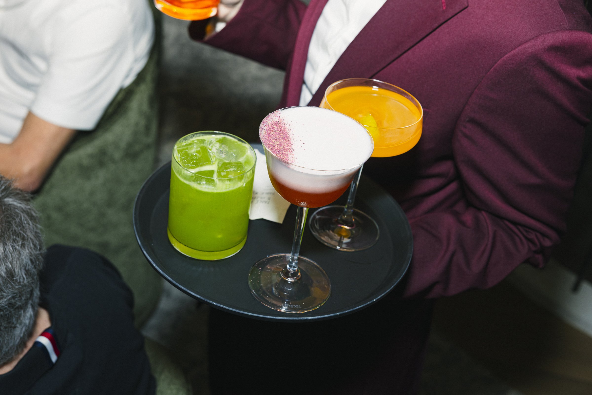 Plateau de cocktails colorés avec un cocktail vert, un cocktail blanc avec déco rose, et un cocktail orange, porté par une personne en veste bordeaux.
