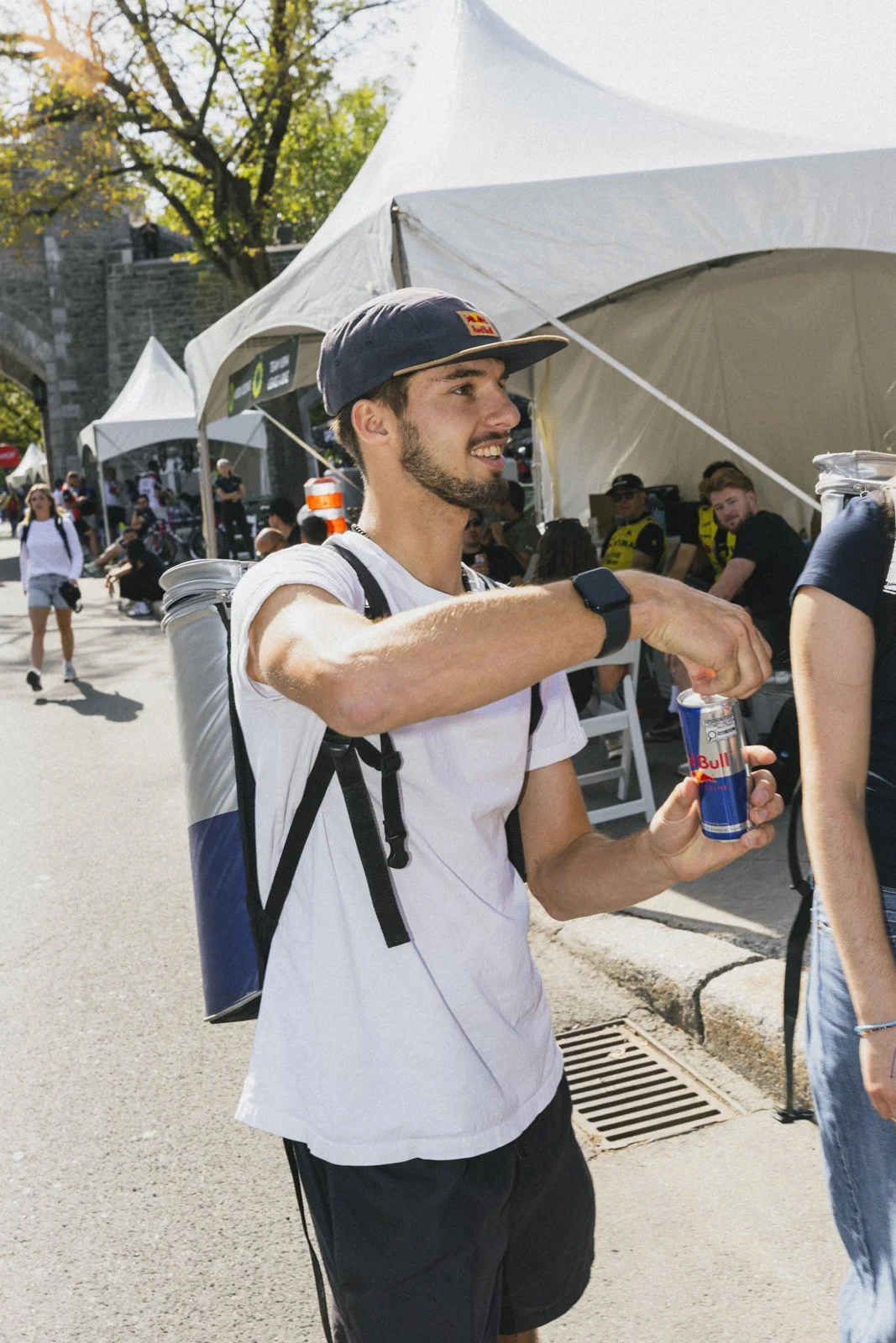Un homme portant une casquette et un t-shirt blanc, tenant une canette Red Bull, souriant lors d'un événement en plein air avec des tentes et d'autres personnes en arrière-plan.