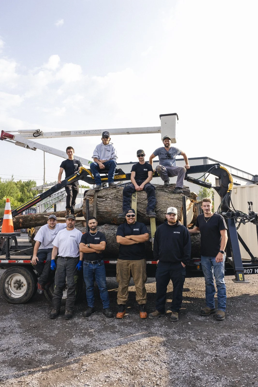 Groupe de dix hommes posant devant un camion avec des troncs d'arbres, certains debout et d'autres assis ou assis sur les troncs, sous un ciel partiellement nuageux.