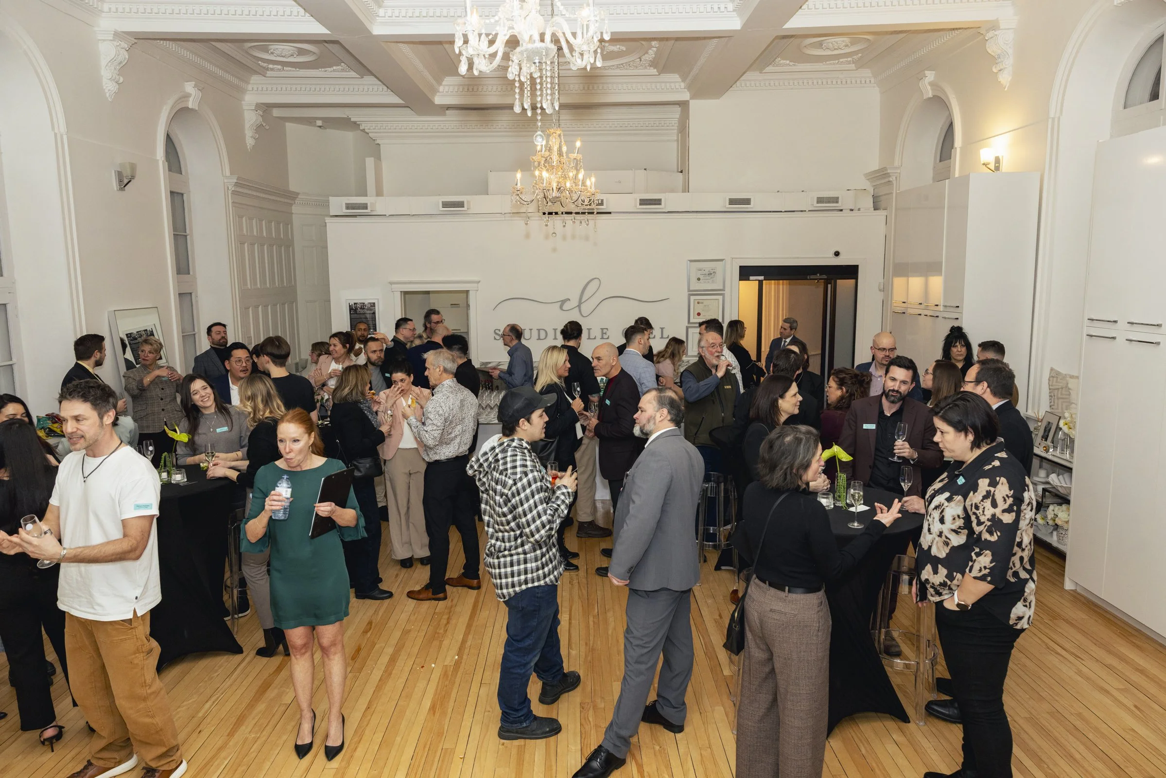 Une réunion ou un événement social avec plusieurs personnes dans une grande salle élégante, discutant, tenant des verres, et socialisant sous un éclairage avec des lustres. La pièce a un style classique avec des murs blancs et des fenêtres arrondies.