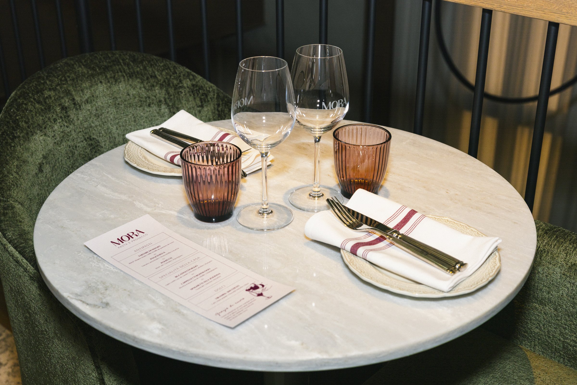Une table ronde en marbre avec des verres à vin, des verres en verre teinté marron, des fourchettes, des assiettes, des serviettes pliées avec une rayure rouge, et un menu, dans un restaurant élégamment décoré.