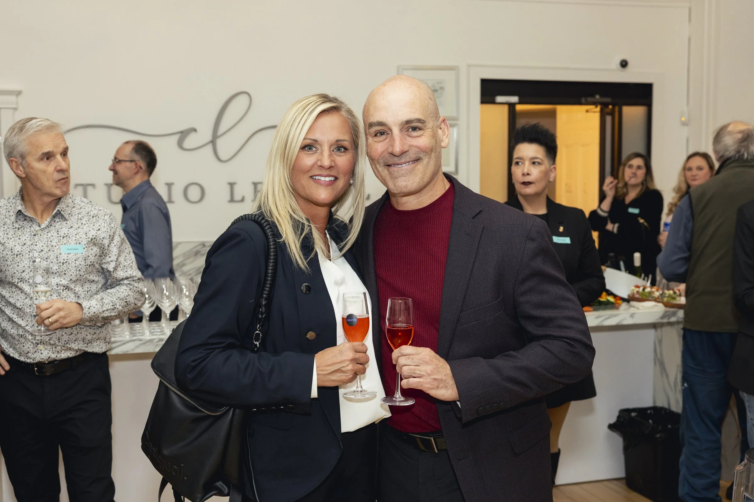 Deux personnes souriantes tenant des flûtes de champagne lors d'une réception, en arrière-plan d'autres invités et un buffet.