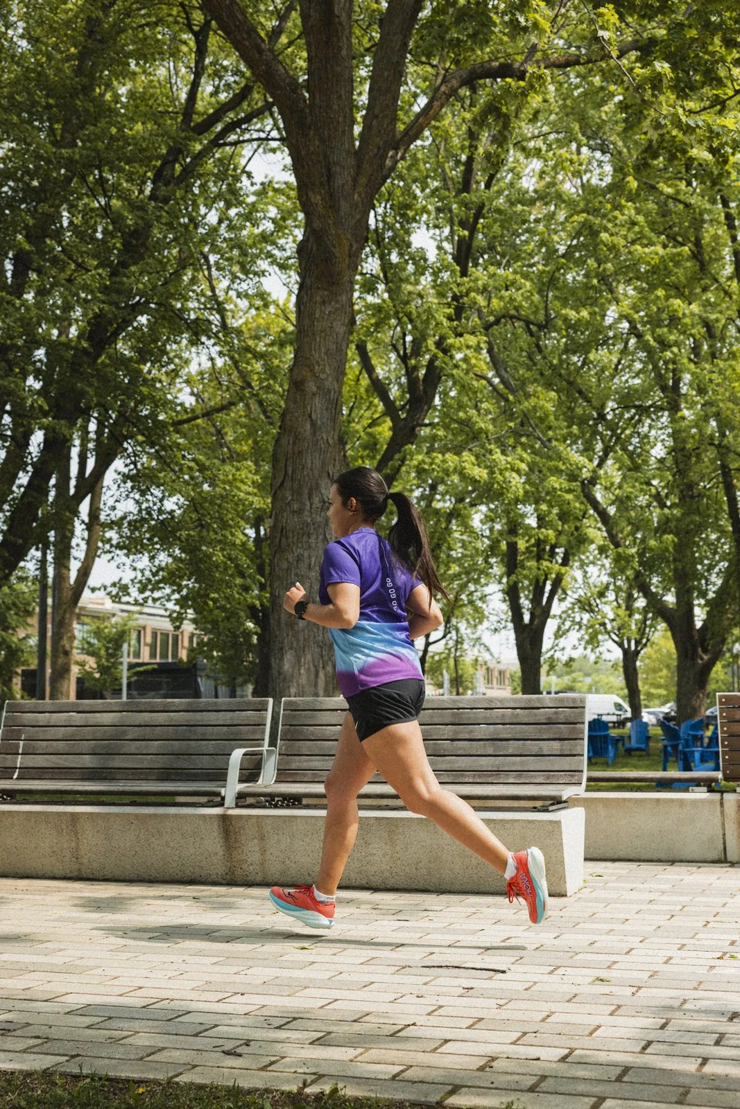 Une femme court dans un parc sous un ciel ensoleillé, entourée d'arbres verdoyants, portant un t-shirt violet, un short noir et des chaussures de course roses