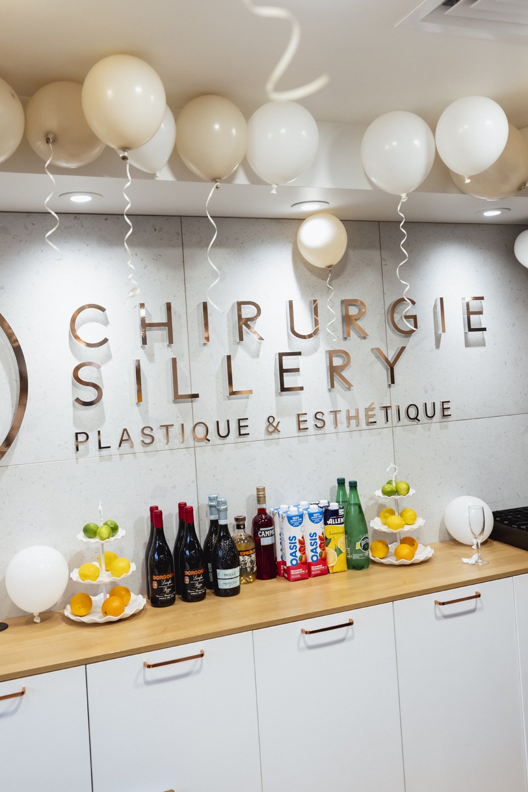 Décoration pour une célébration à la clinique de chirurgie plastique avec des ballons blancs, des bouteilles, des jus, et des desserts aux fruits.
