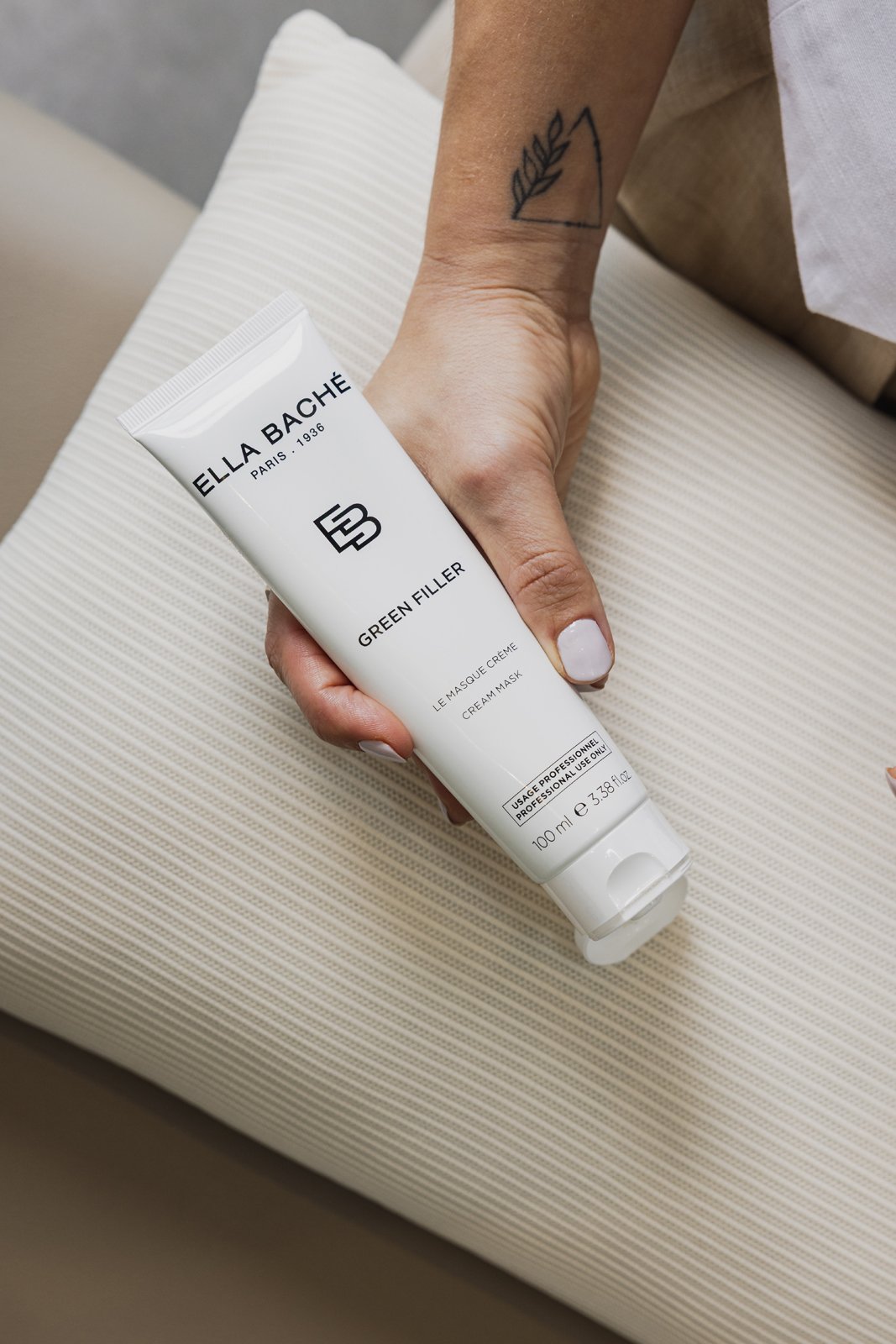 Main d'une personne tenant un tube de masque facial Ella Baché Green Filler, posé sur un tissu beige rayé.