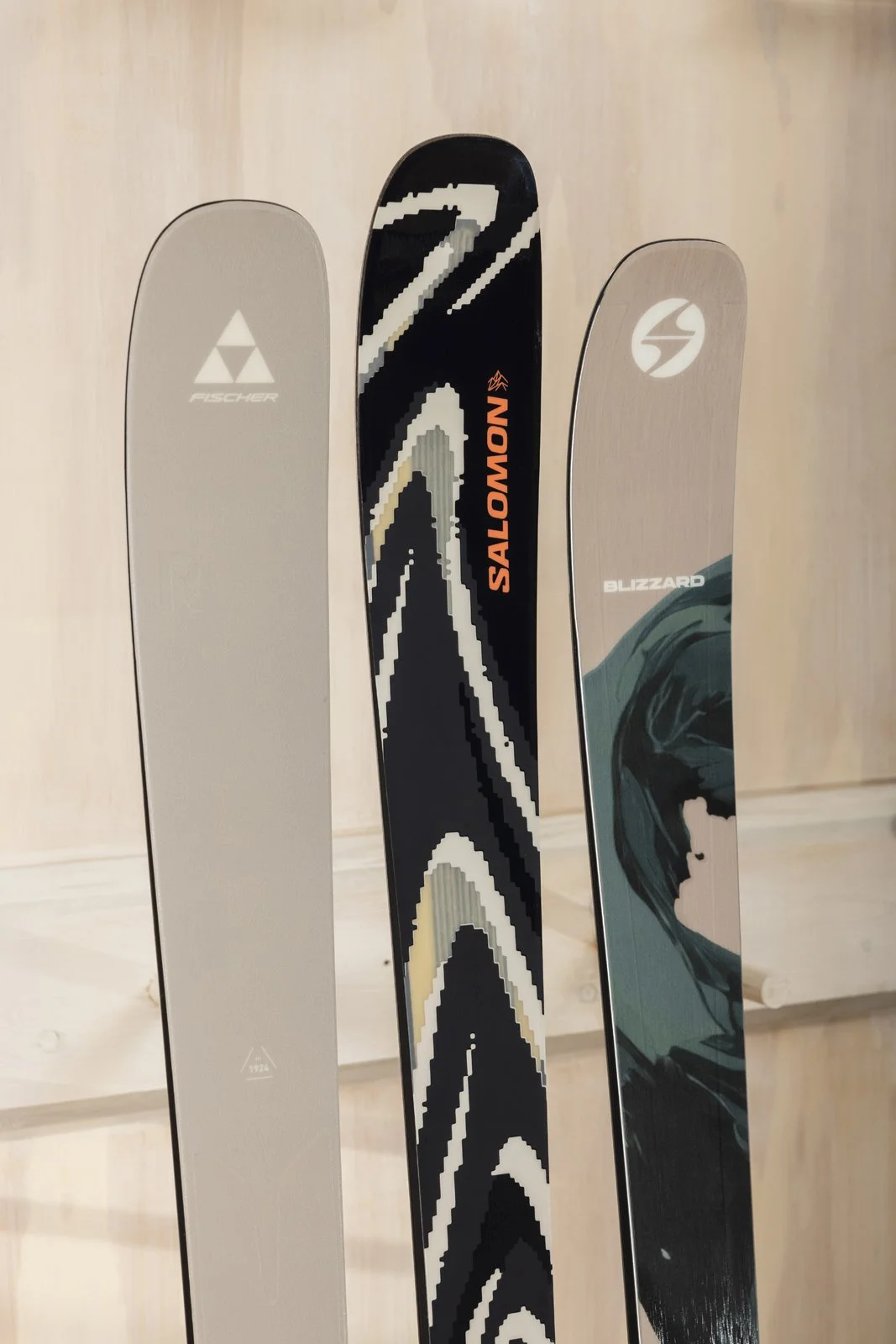 Trois skis sont alignés verticalement. Le premier à gauche est beige avec le logo 'Fischer'. Le milieu est noir avec des motifs en blanc, gris, et orange, avec le texte 'SALOMON' en orange. Le troisième à droite est blanc avec un logo circulaire et u