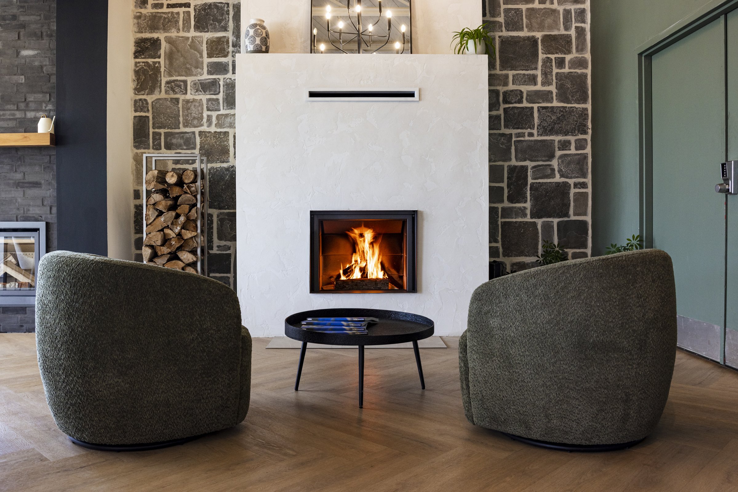 Salon avec cheminée moderne allumée, deux fauteuils verts, et une table ronde noire, dans une pièce avec murs en pierre et sol en bois.