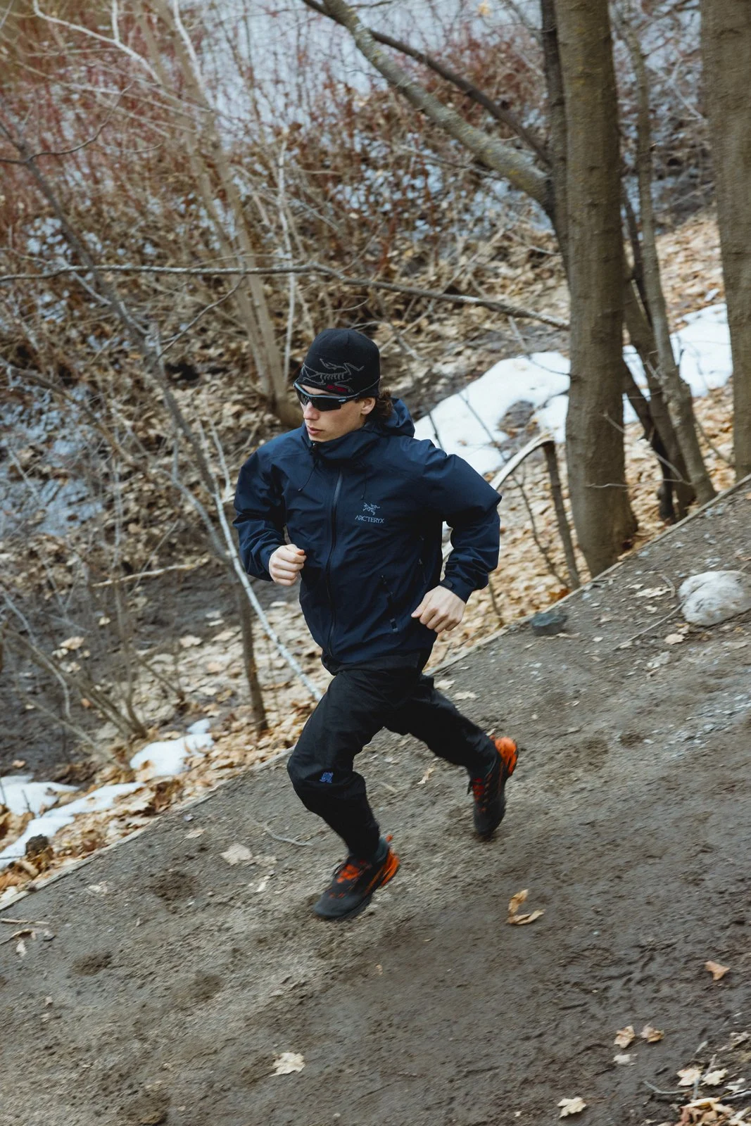 Un homme en vêtements de course en extérieur, portant une veste noire, un bonnet noir et des lunettes de soleil, court sur un sentier en terre dans une forêt avec des feuilles mortes et quelques restes de neige.