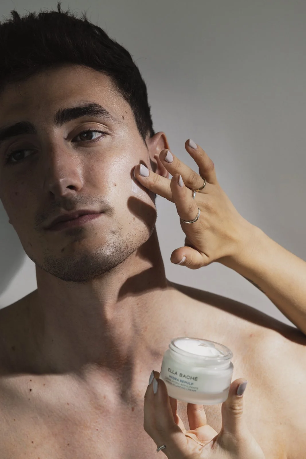 Un homme jeune avec des cheveux courts et des taches de rousseur appliquant de la crème sur sa joue, tenant un pot de crème de visage Ella Baché, avec une main féminine avec plusieurs bagues.