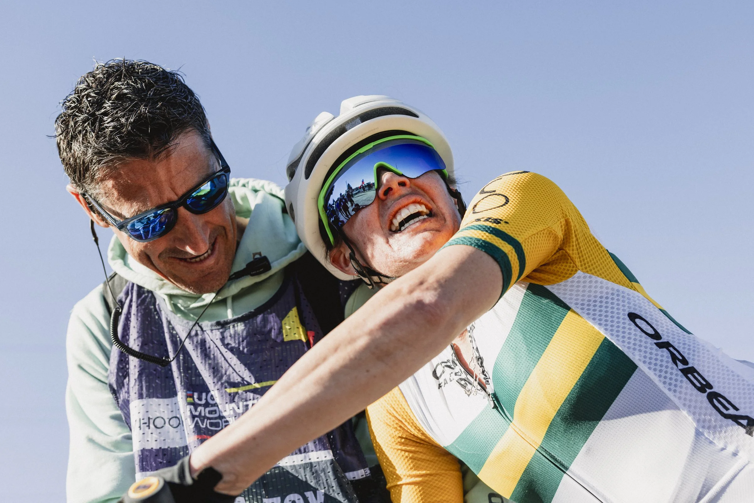 Un homme en tenue de cycliste jaune et blanc, portant un casque et des lunettes, semble avoir chuté ou été aidé après une chute tandis qu'une autre personne en veste avec un logo, portant des lunettes de soleil, l'aide avec un sourire.