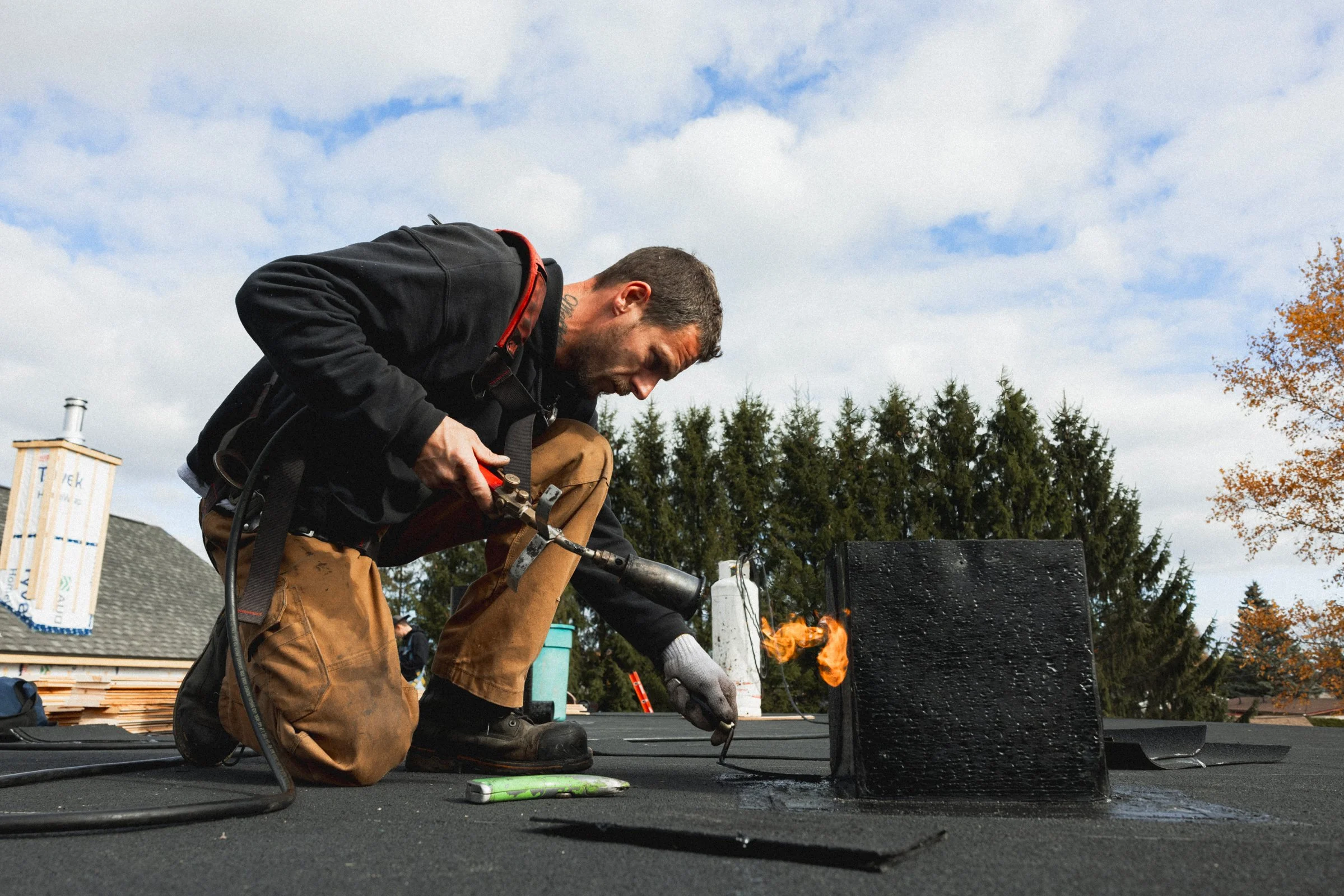 Un homme en train de faire une soudure sur un toit, avec une flamme orange visible, portant des vêtements de travail et une paire de gants de protection, avec un ciel nuageux en arrière-plan.