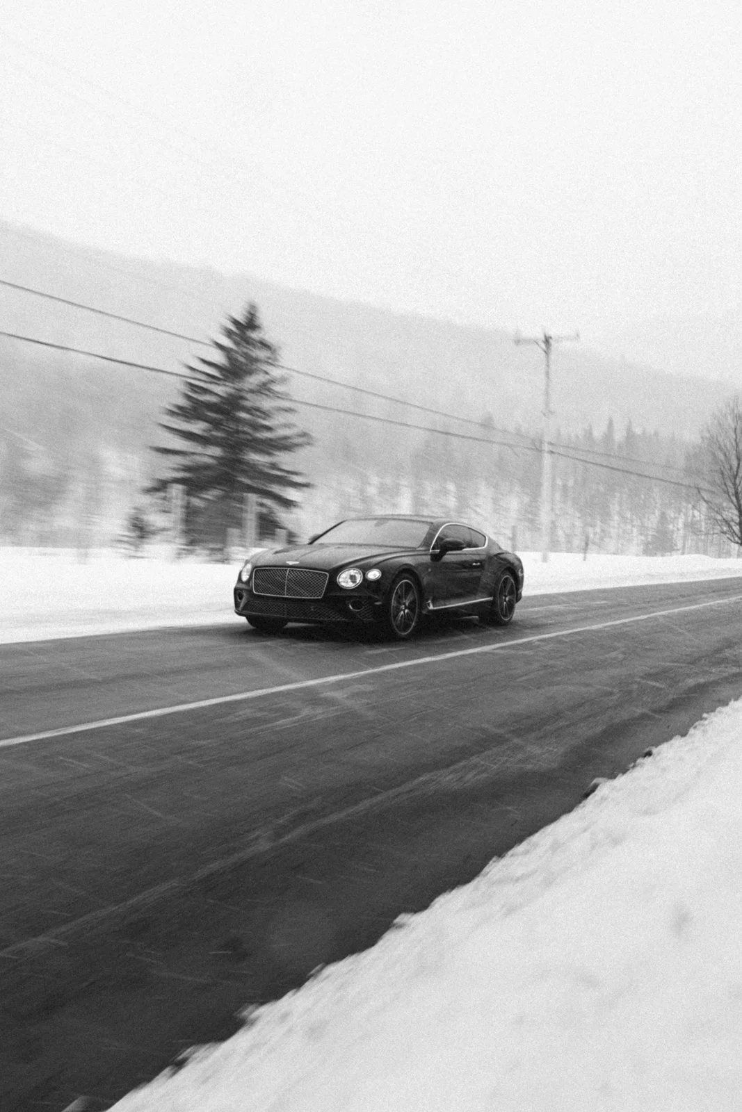 Voiture noire roulant sur une route enneigée avec des arbres et des montagnes en arrière-plan dans un paysage hivernal, en noir et blanc.