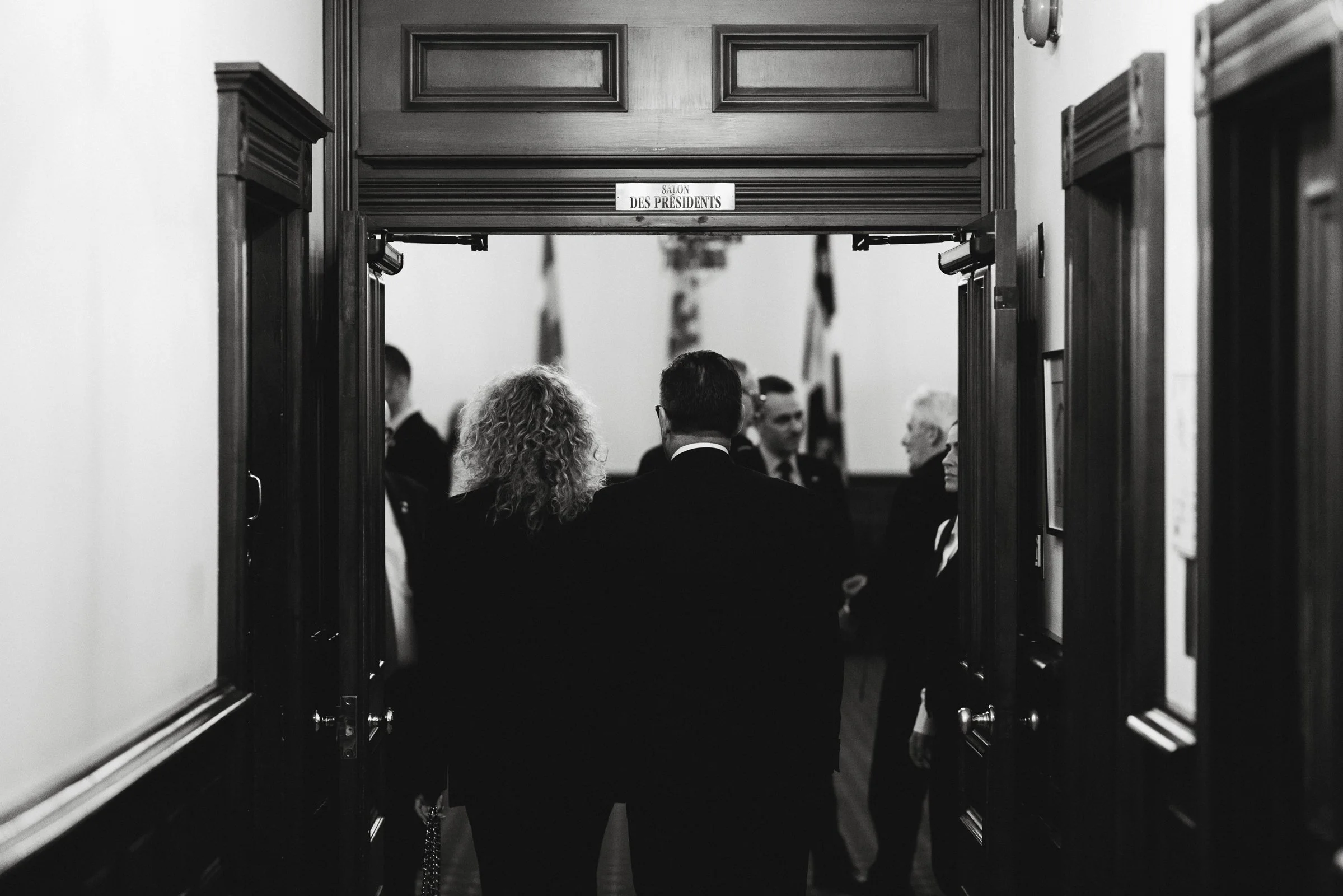 Personnes en costume dans un couloir menant à une salle, vue de dos, avec un panneau indiquant 'Salon des Présidents'