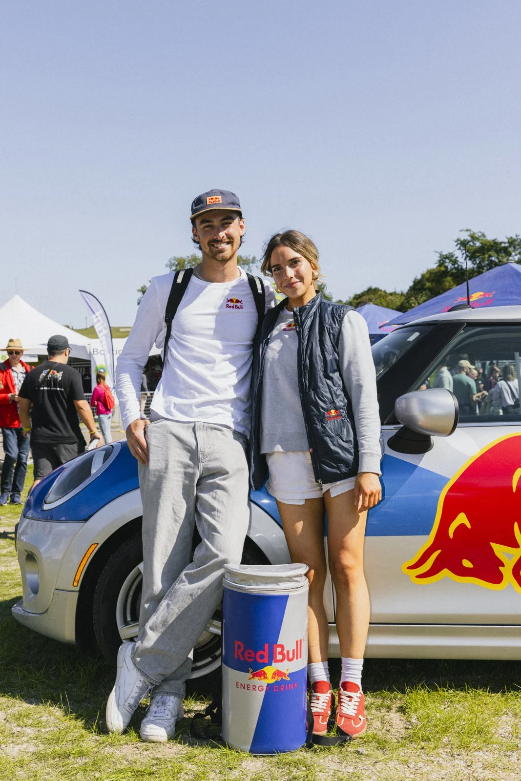 Deux jeunes adultes posent devant une voiture sponsorisée Red Bull lors d'un événement en plein air. L'homme porte un t-shirt blanc, un sac à dos, un chapeau et des chaussures blanches. La femme porte un sweat gris, un gilet noir, des shorts blancs e