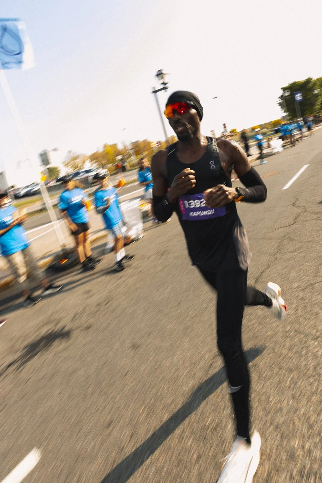 Un coureur à pied en pleine course lors d'une compétition, portant une tenue noire, des lunettes de soleil rouges et une montre sportive, avec des bénévoles en bleu en arrière-plan.