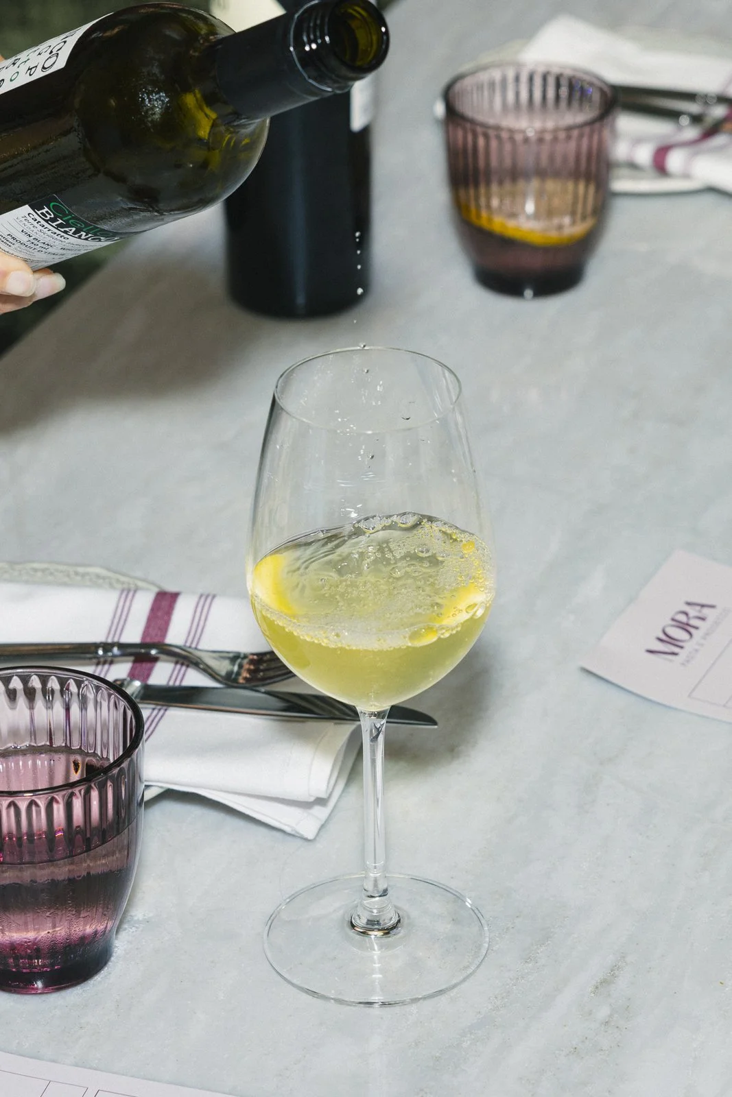 Un verre de vin blanc avec une tranche de citron dans un dineau blanc, sur une table avec des couverts, un verre rose foncé et un menu.