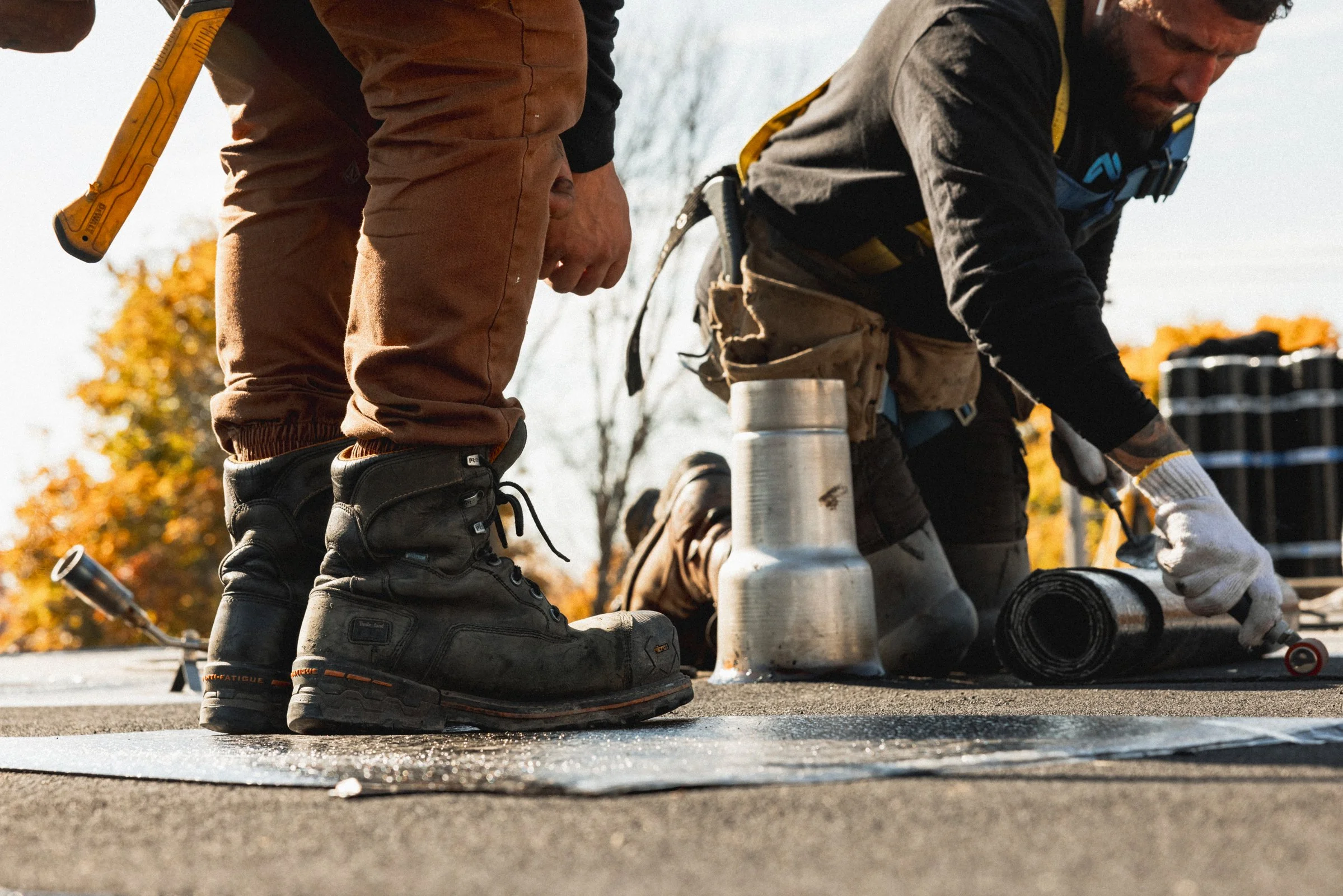 Deux ouvriers travaillent sur la pose d'une nouvelle couche de goudron sur une route en été, avec des arbres aux feuilles orange en arrière-plan. Ils portent des chaussures de sécurité, et un des deux est à genoux, manipulant un rouleau compresseur.