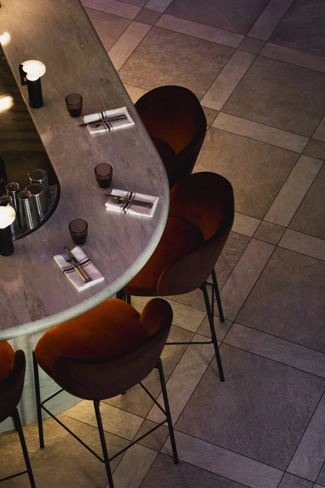 Table de restaurant ronde en marbre avec trois chaises en bois et métal, nappes, couverts, verres, et assiettes, vue depuis le dessus.