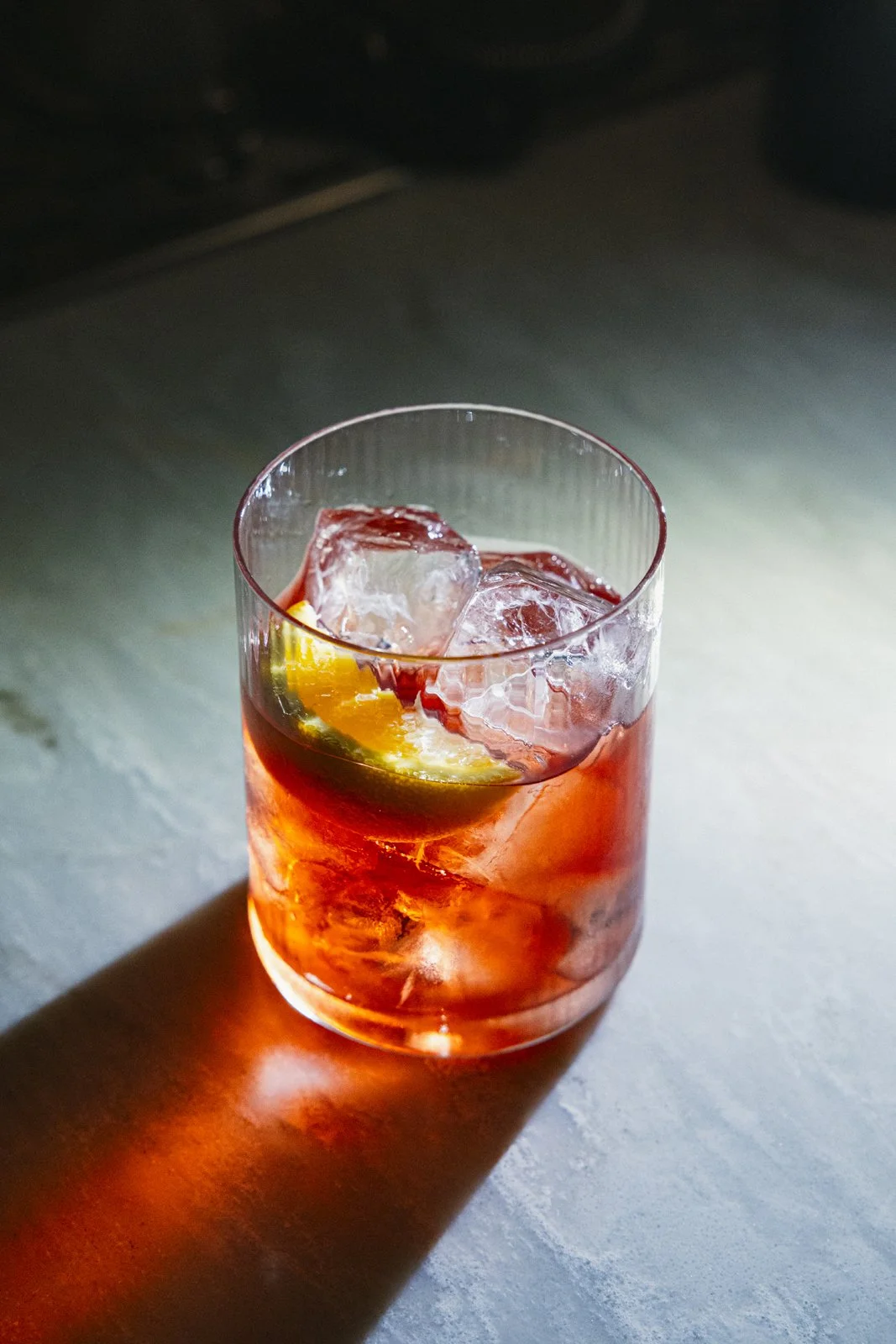 Verre de cocktail rouge avec des cubes de glace et une tranche de citron, éclairé par la lumière créant une ombre colorée au sol.