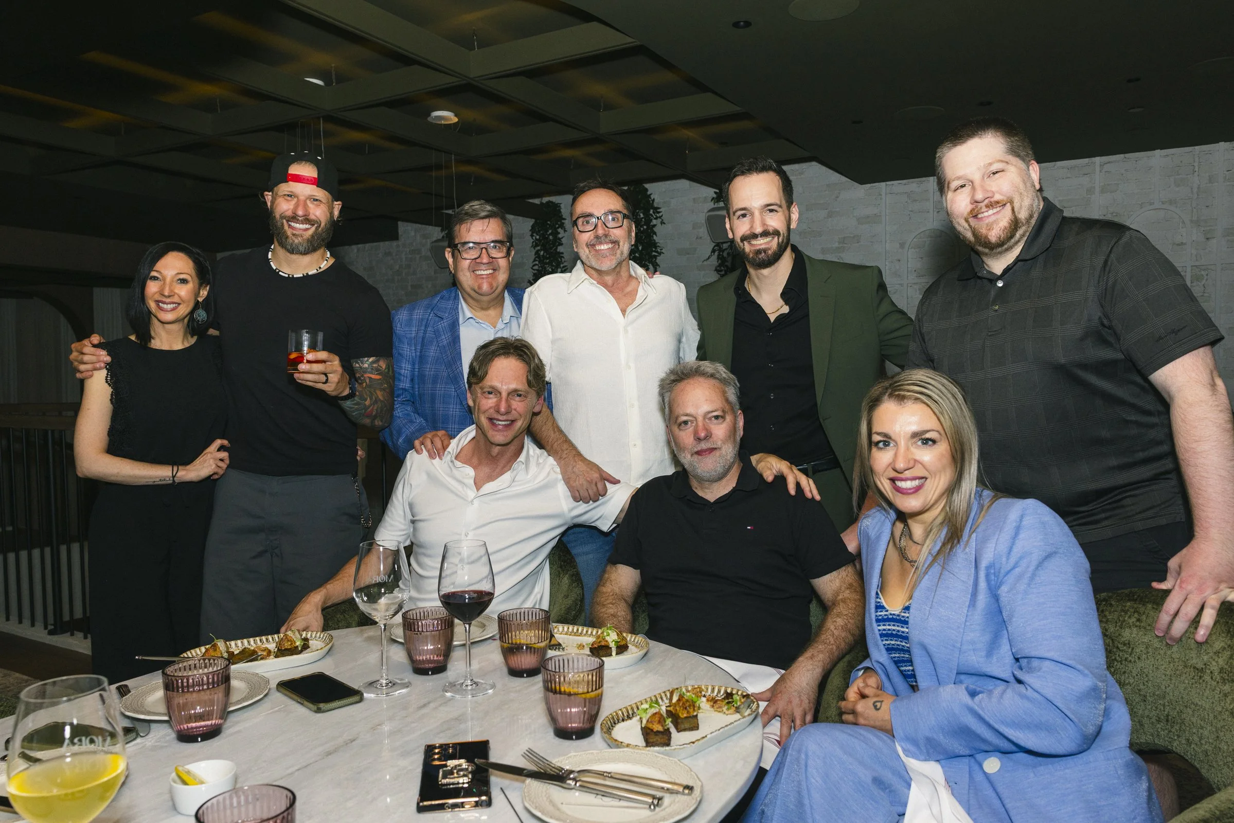 Groupe de dix personnes souriantes lors d'un dîner, assises autour d'une table avec de la nourriture et des boissons, dans un restaurant ou une salle de réception élégante.