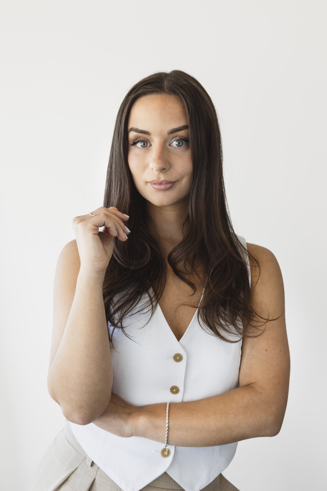 Jeune femme aux cheveux bruns, yeux bleus, portant un top blanc avec boutons, posant contre un fond blanc.