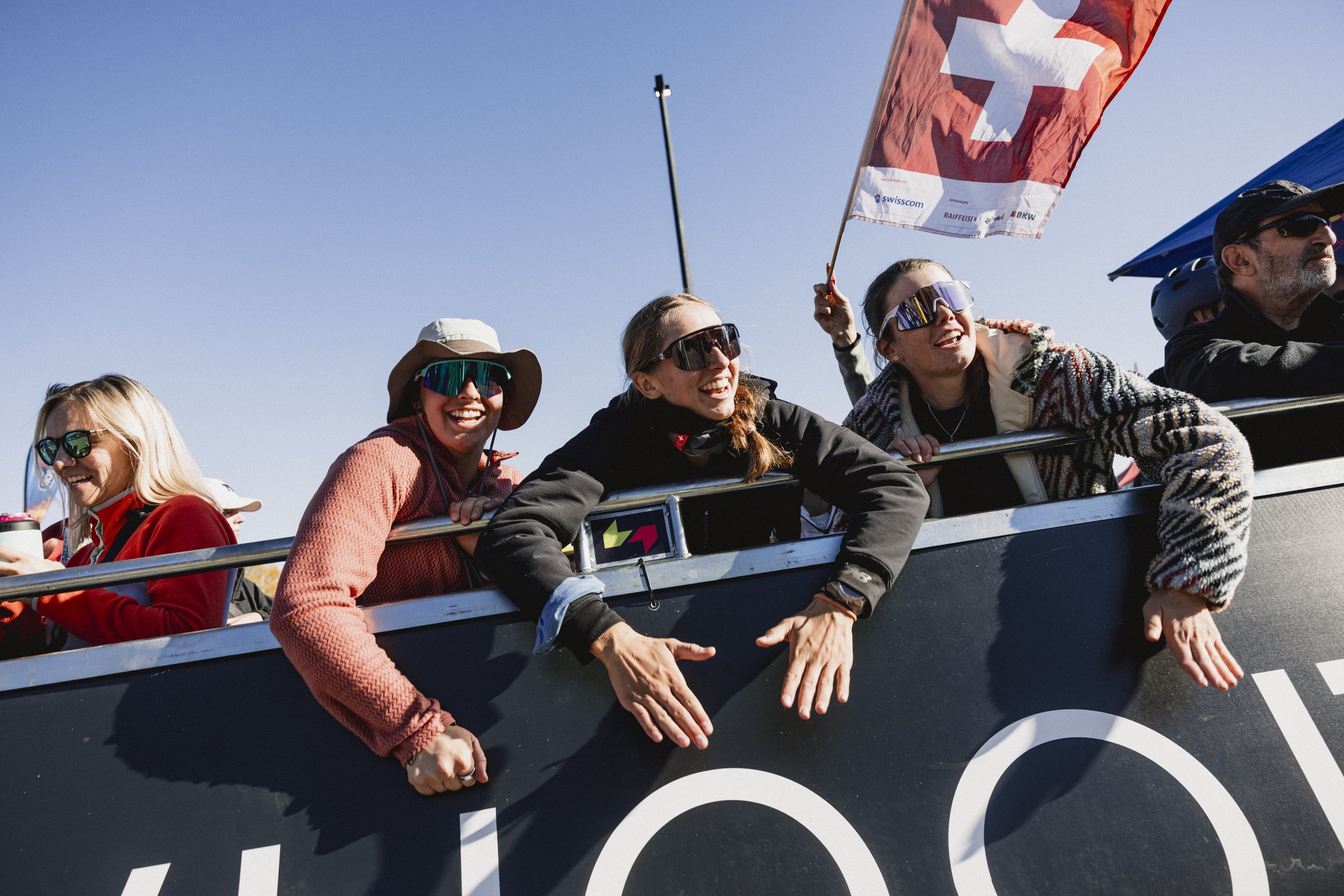 Groupe de personnes souriantes en tenue de sport, portant des lunettes de soleil, sur un bateau durant une compétition de voile, avec un drapeau suisse flottant en arrière-plan.