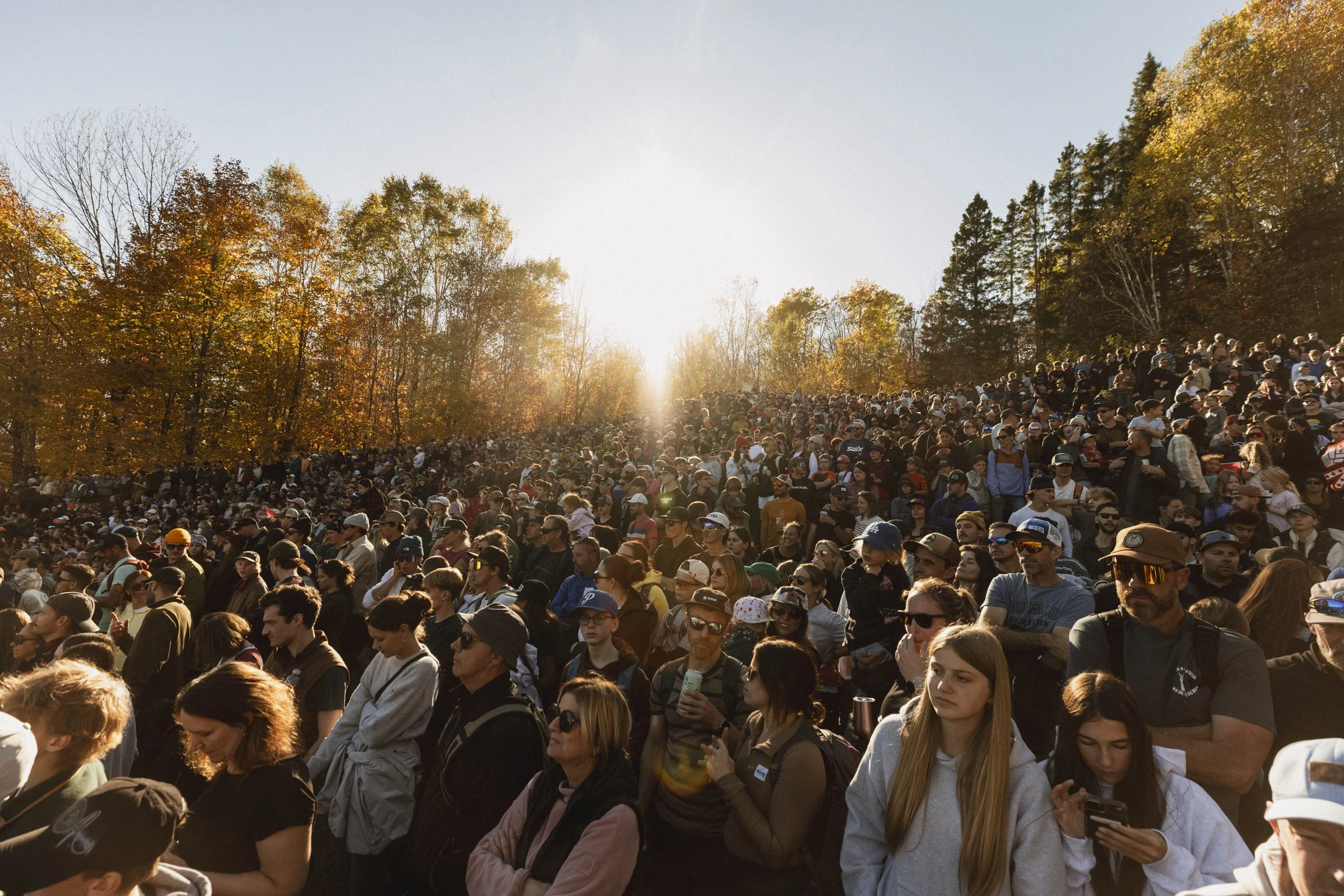 Une grande foule de personnes assistent à un événement en plein air lors d'une journée ensoleillée d'automne, entourée d'arbres aux feuillages colorés.
