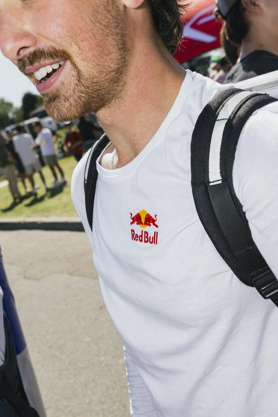 Un homme souriant porte un t-shirt blanc avec le logo Red Bull. Il porte un sac à dos noir. La scène semble se dérouler à l'extérieur avec d'autres personnes en arrière-plan.