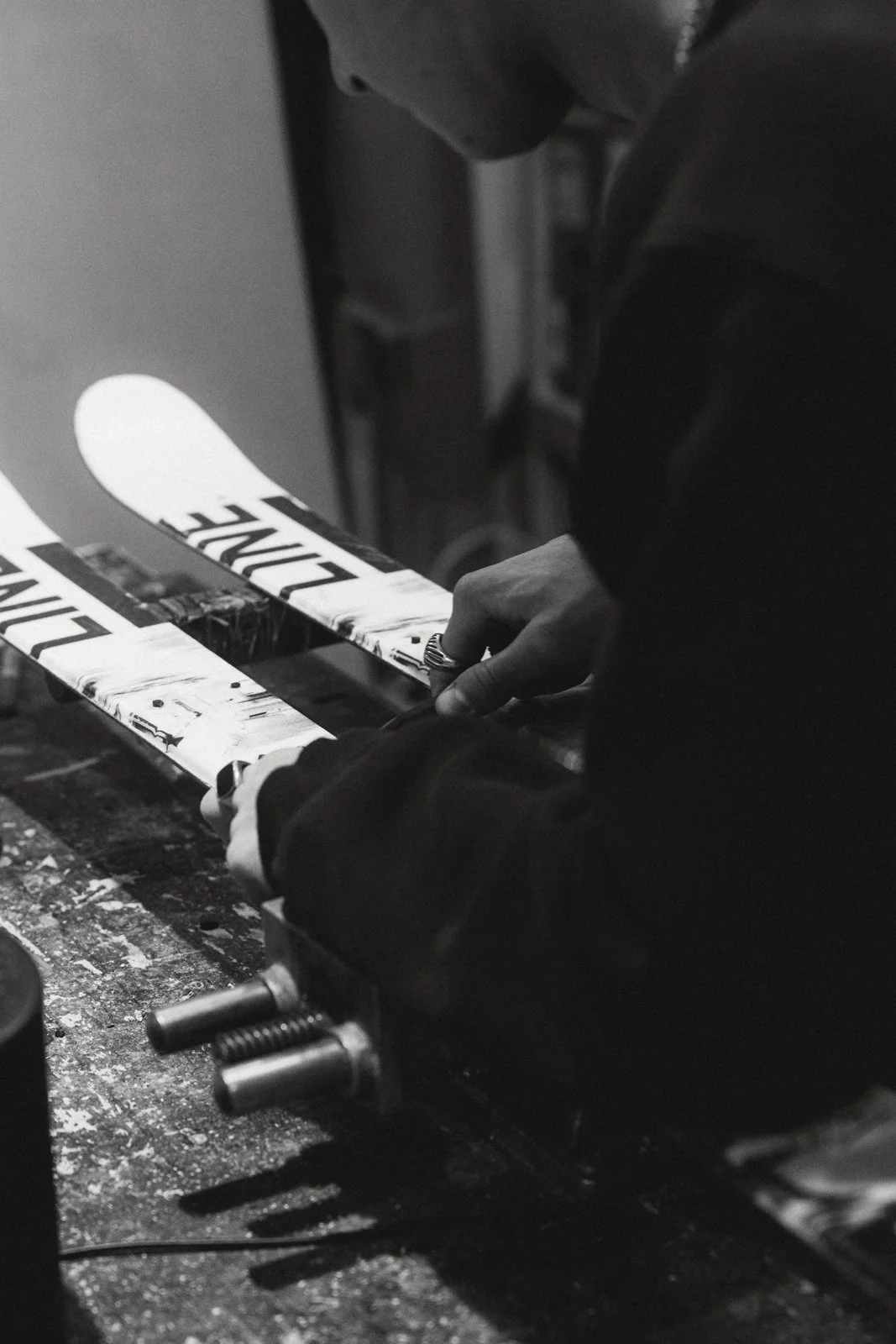 Une personne ajuste une paire de skis à l'aide d'outils dans un atelier. Les skis portent le mot 'ZIPLINE'. L'image est en noir et blanc.