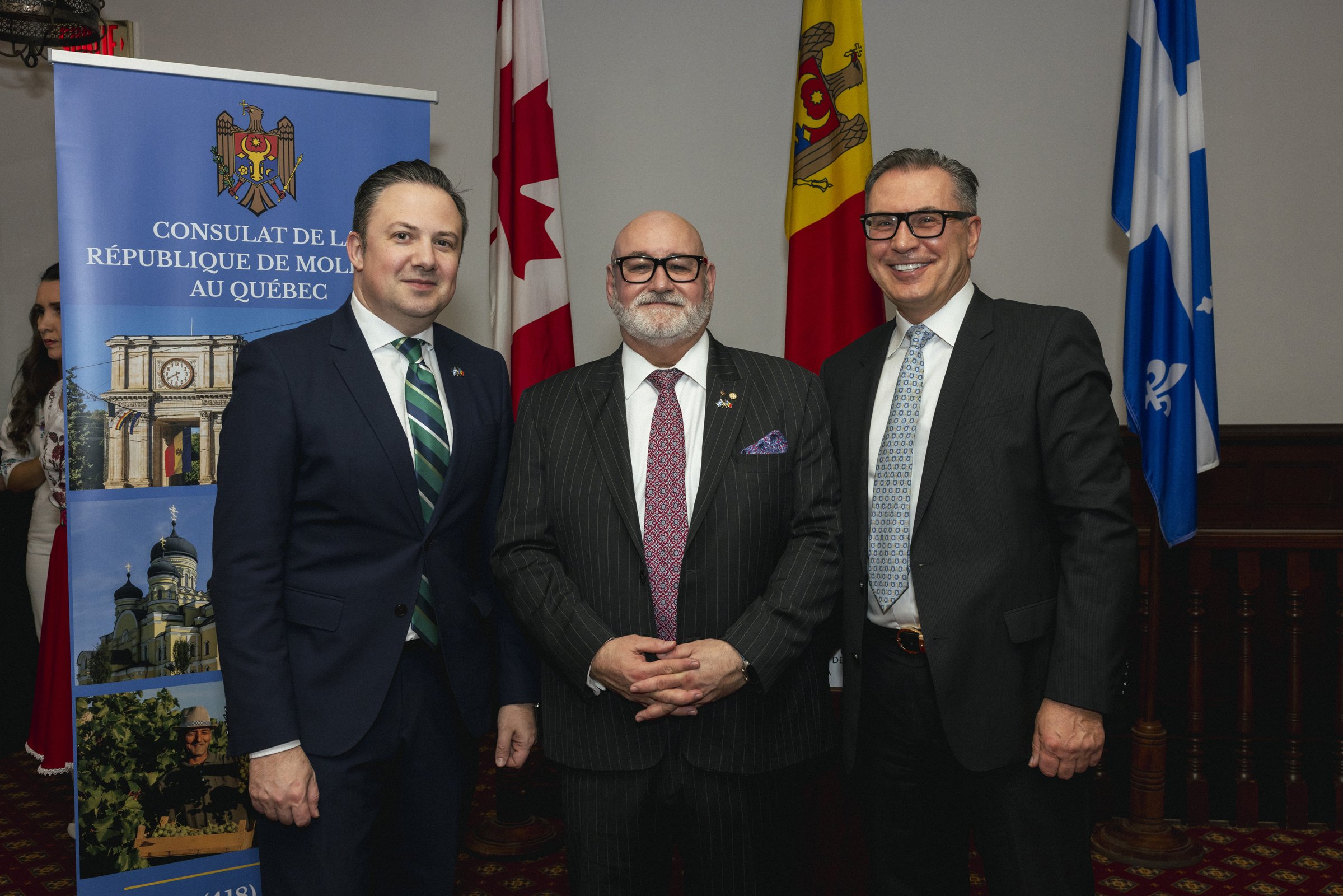 Trois hommes en costumes posent pour une photo devant une bannière et des drapeaux lors d'une réunion à l'ambassade de Moldavie au Québec.